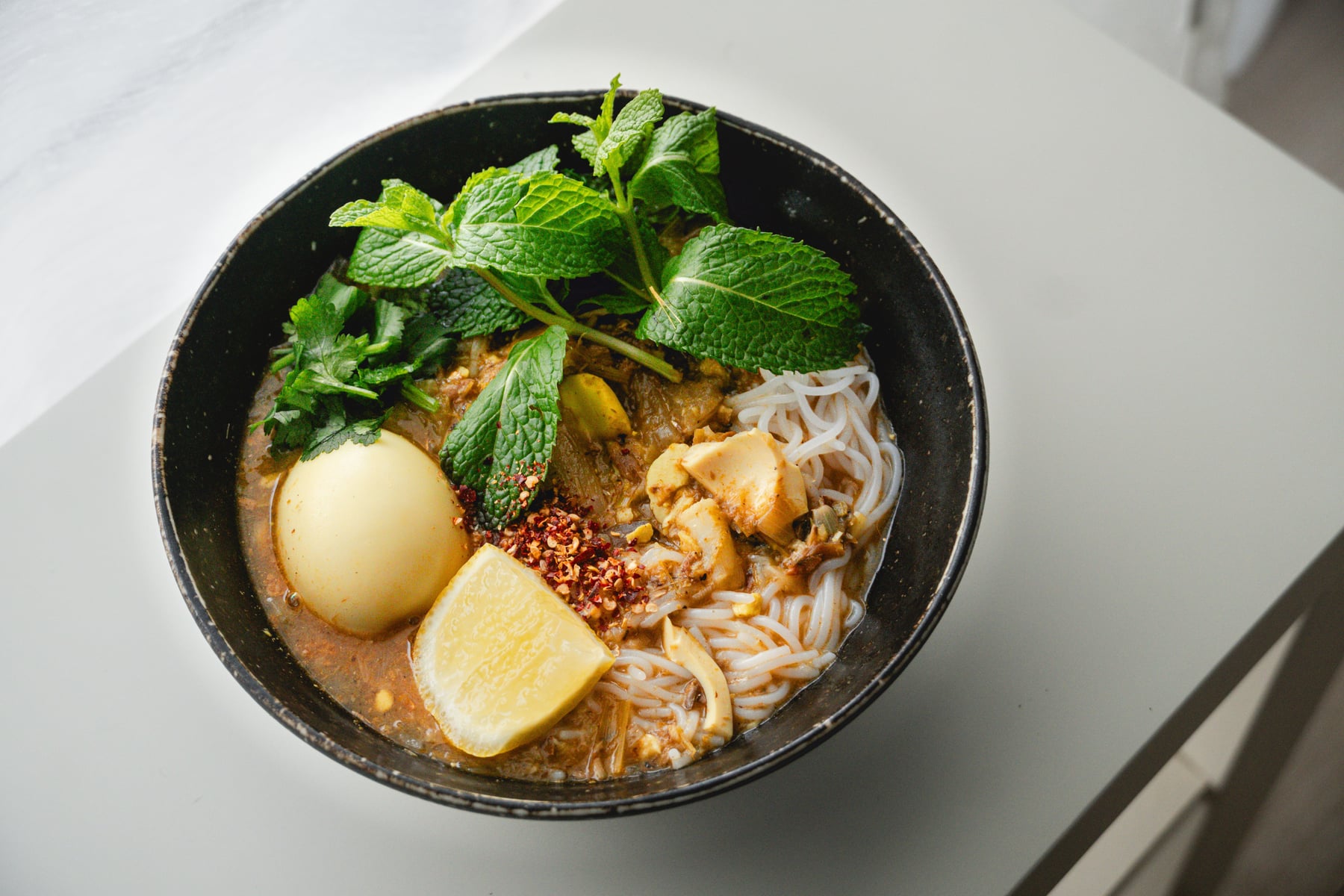 A bowl of Burmese noodle soup topped with a boiled egg, lime, mint and coriander, chilli flakes scattered over the broth.