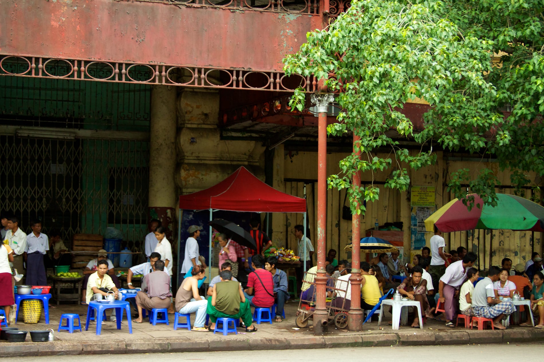 Groups of people sit on low plastic stools at street-side teashop tables in downtown Yangon, shaded by a tree and colonial-era buildings.