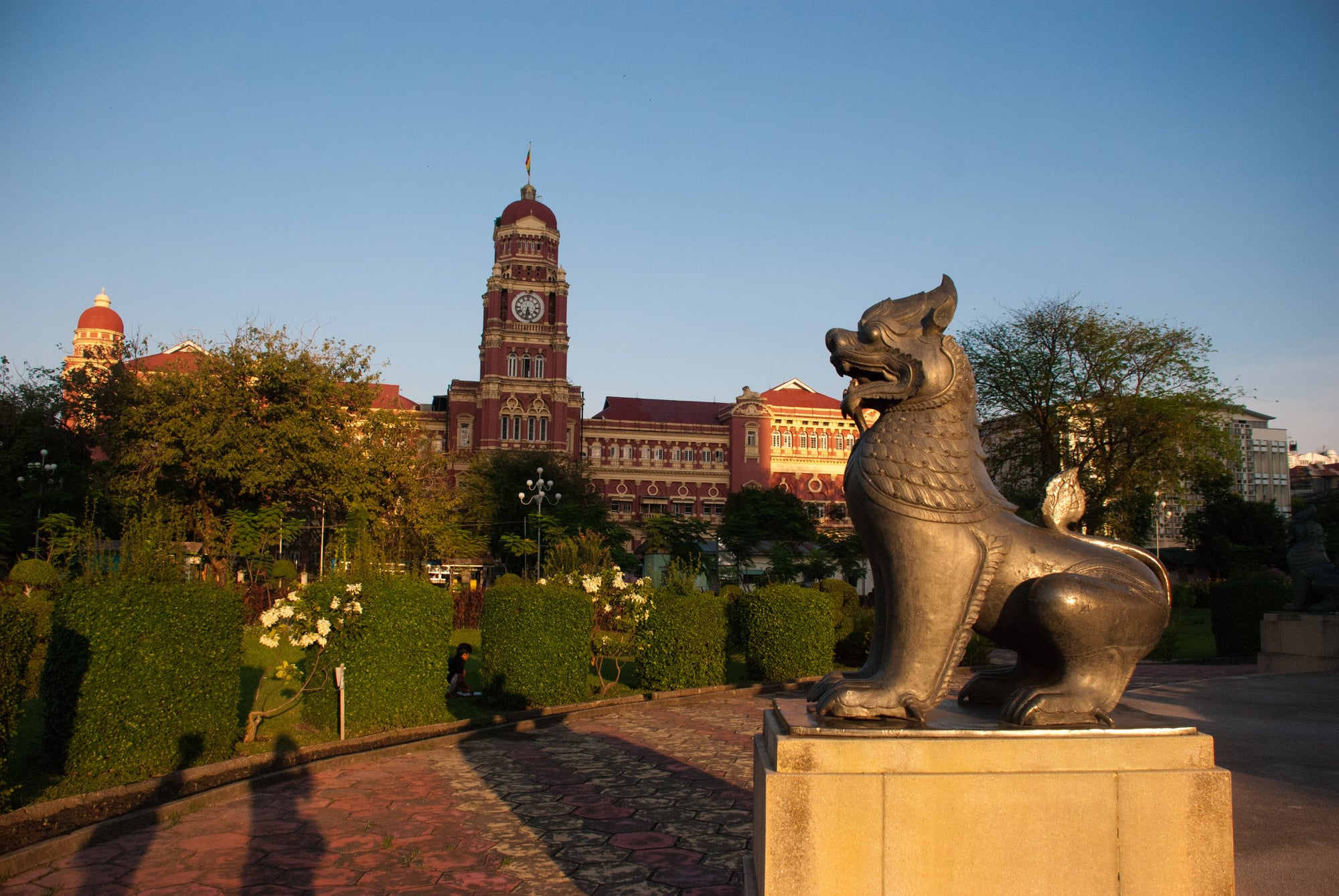 A bronze chinthe statue stands in Mahabandula Park at golden hour, with the red-brick clock tower of the former High Court behind it.