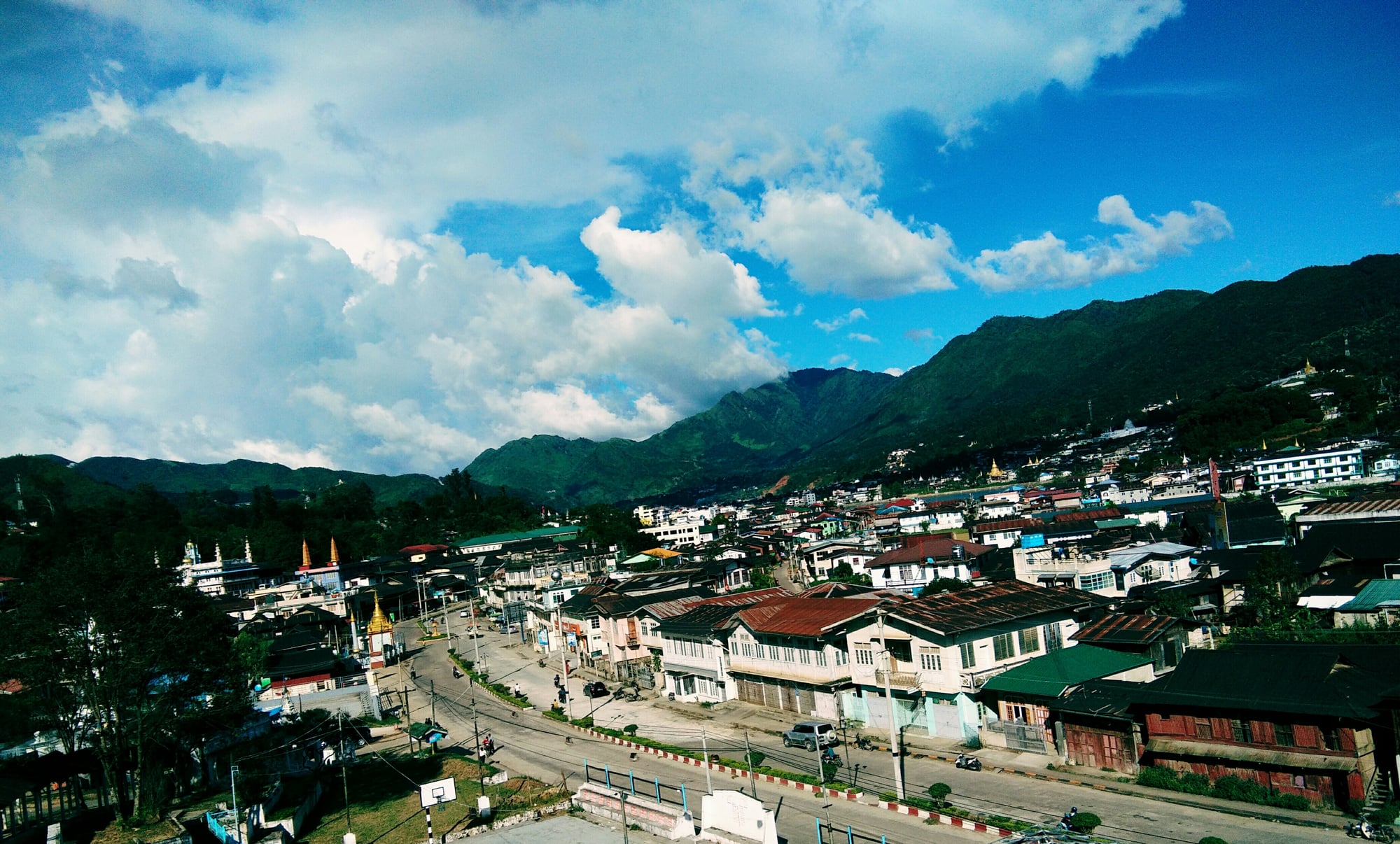 A wide view of Mogok township in Myanmar, its buildings and golden pagoda spread across a valley between forested hills.