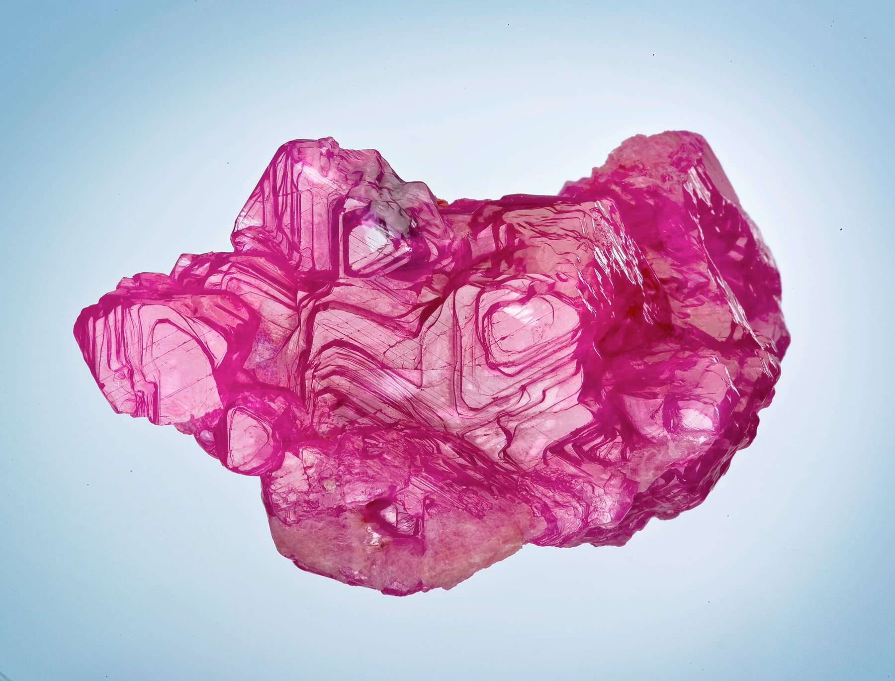 A rough ruby crystal from Mogok, Myanmar, glowing deep pink-red against a pale background.