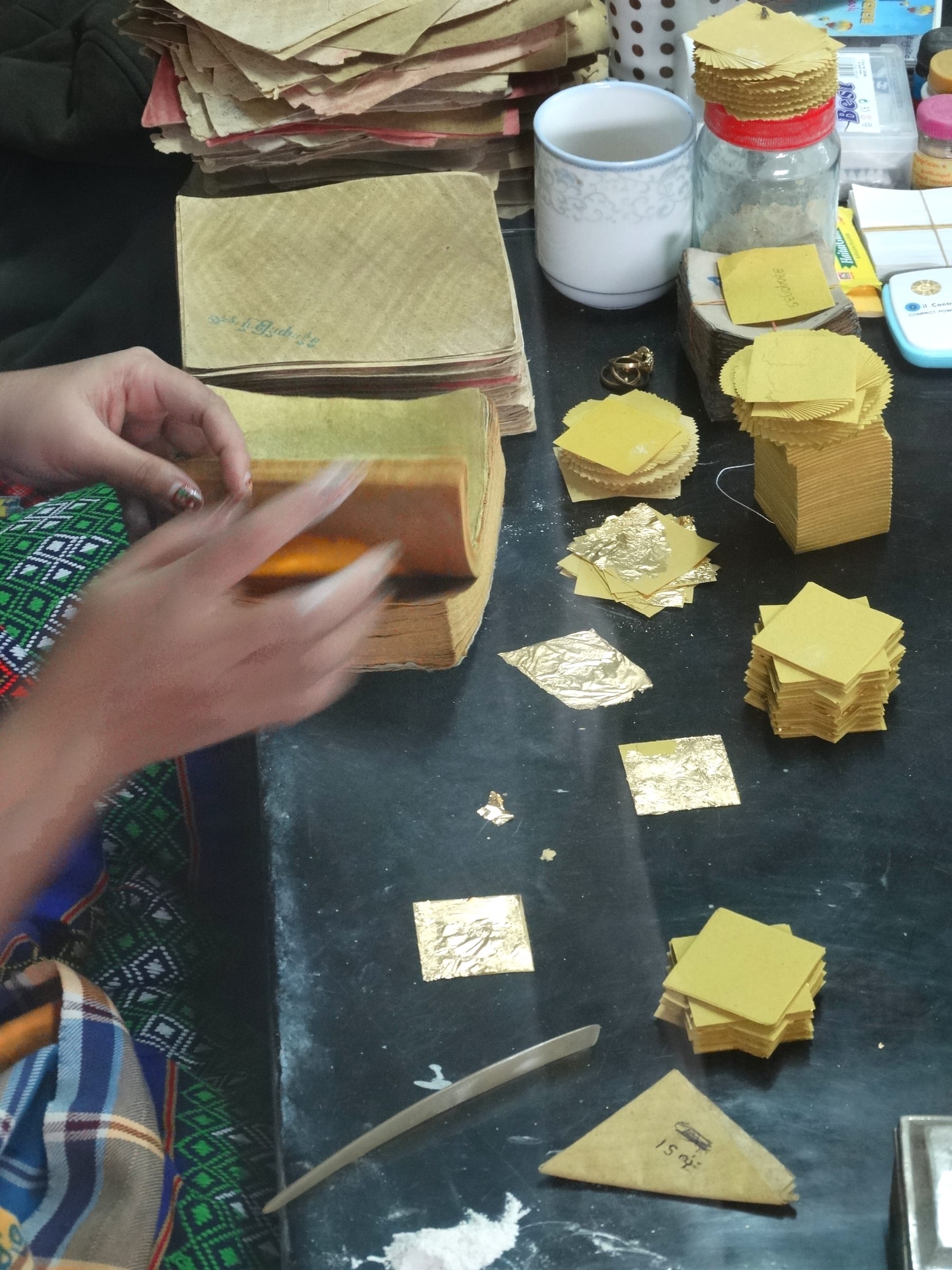 Hands cutting and sorting delicate squares of beaten gold leaf onto small paper backings at a workshop table.