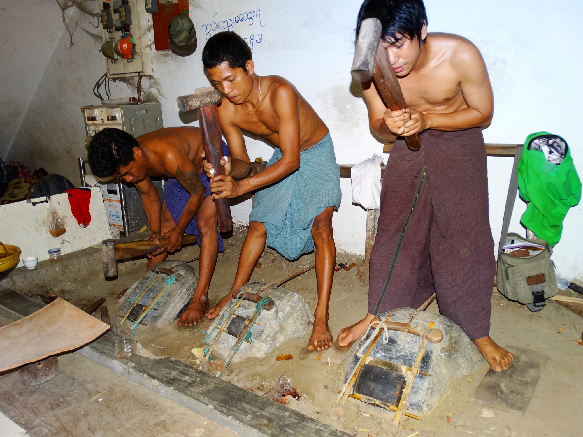 Three shirtless workers in longyi rhythmically hammer stone-wrapped bundles of gold leaf at a Mandalay workshop.