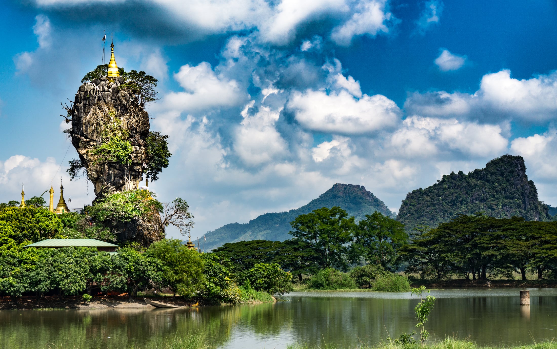 A golden pagoda crowns a narrow limestone pinnacle rising from a lake near Hpa-An, with mountains and blue sky behind it.