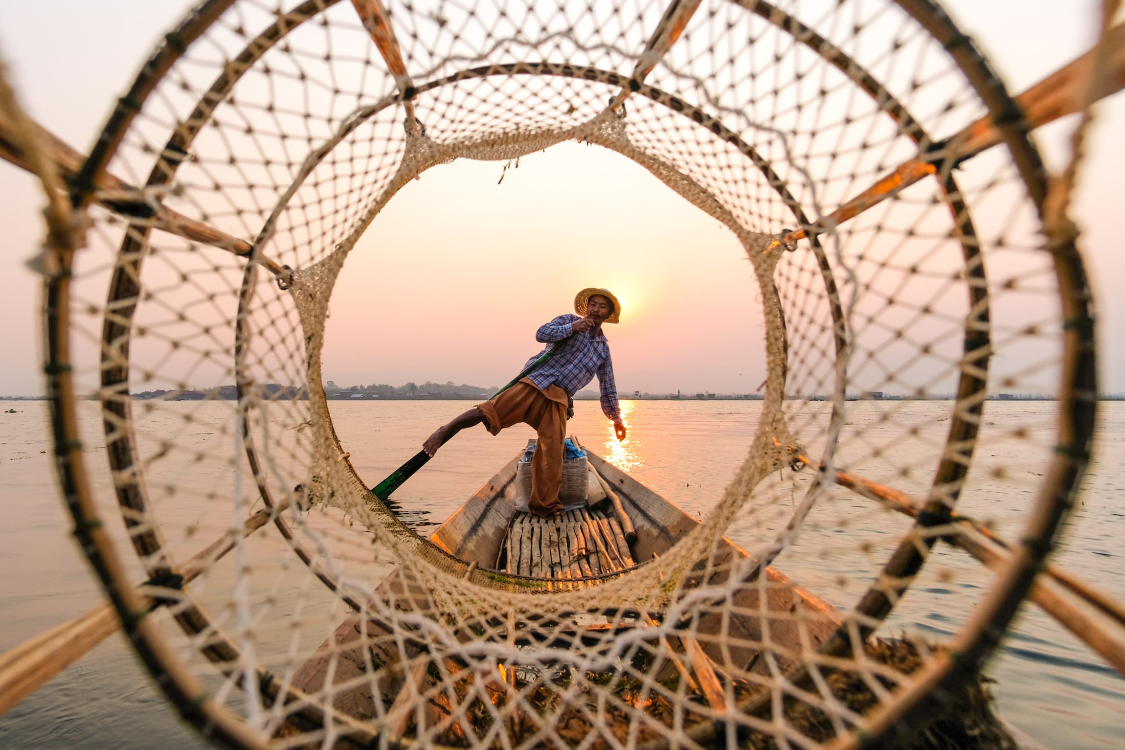 Seen through the circular frame of a conical fishing basket, an Inle Lake fisherman balances on one leg at sunset, rowing with the other.