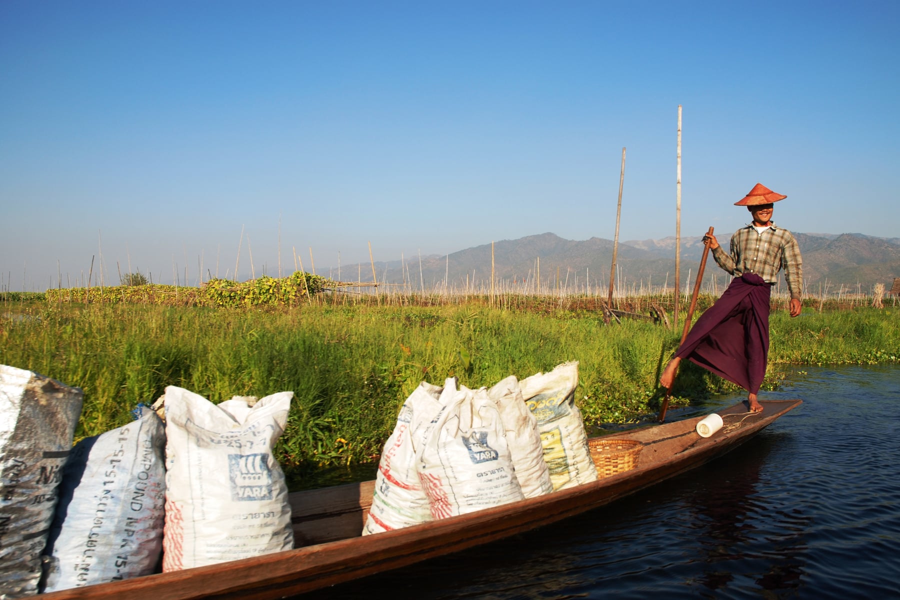 A leg-rowing farmer transports sacks of produce past rows of floating garden beds, with the Shan hills in the distance.