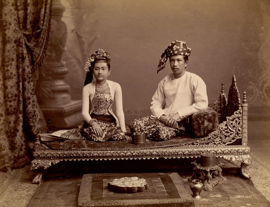 A sepia-toned studio portrait of a Burmese couple in elaborate precolonial court dress, seated on an ornately carved daybed.