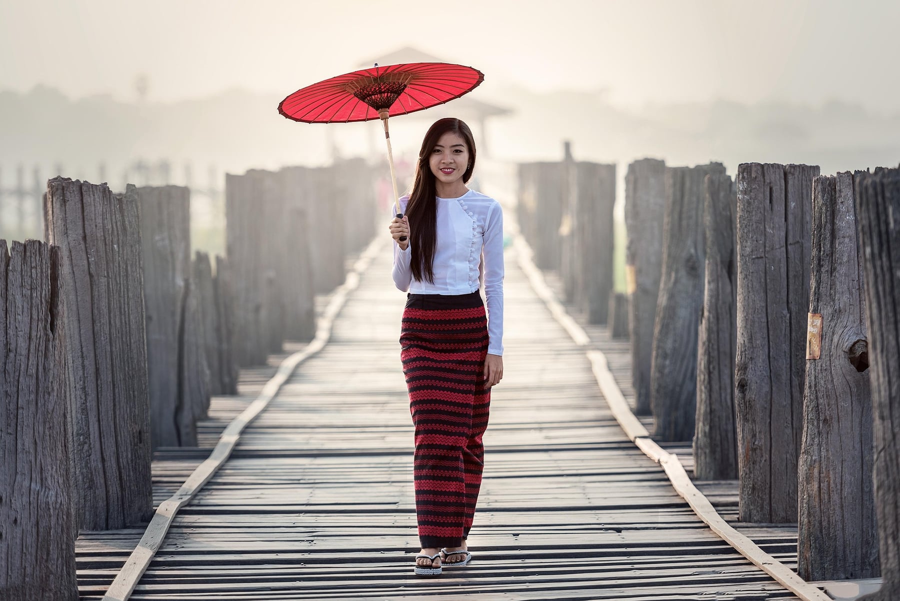 A young woman in a striped red and black htamein holds a red parasol while walking across U Bein Bridge in Mandalay.