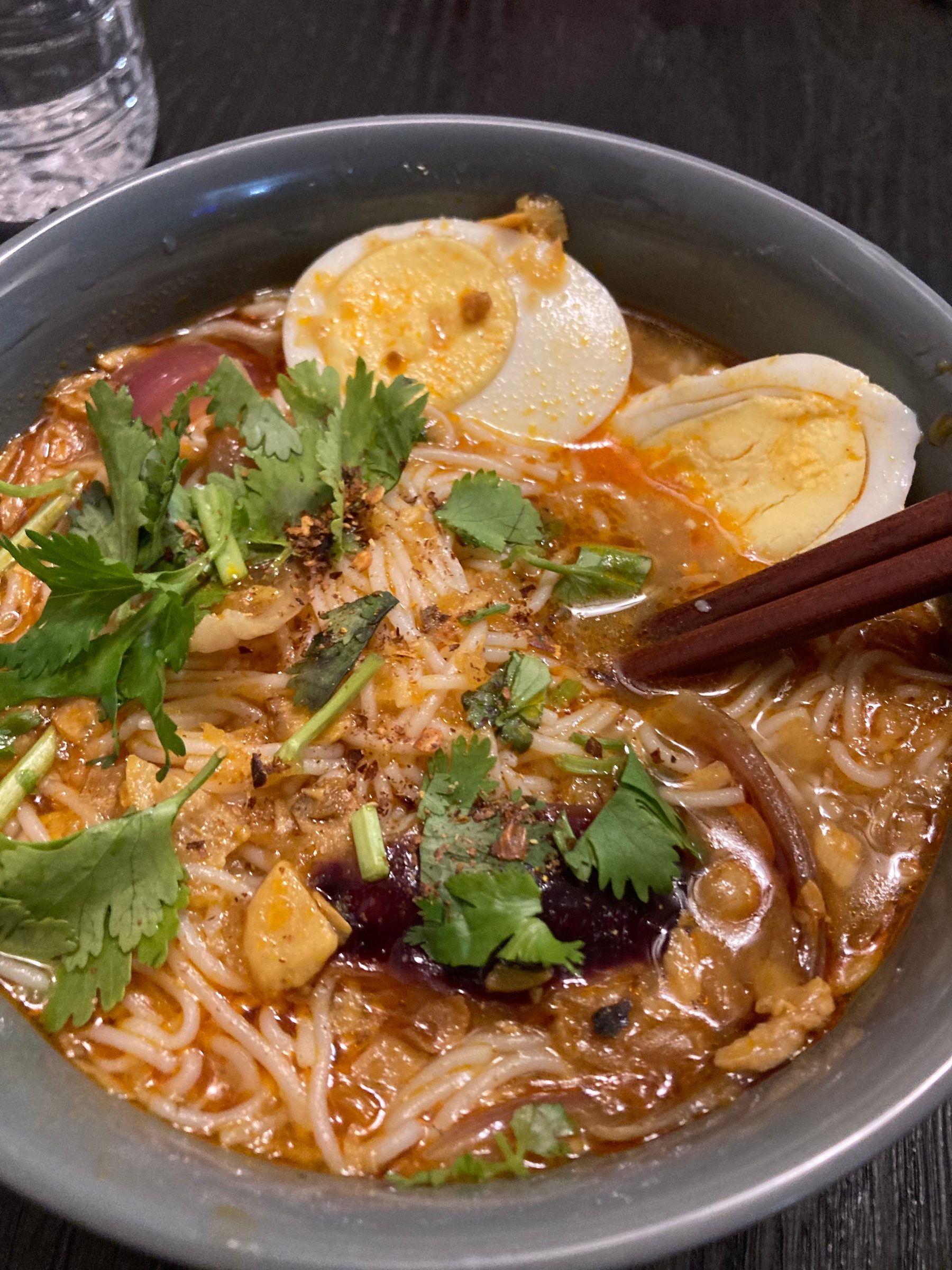 An overhead view of a bowl of mohinga fish noodle soup topped with sliced boiled egg and coriander, served with chopsticks.