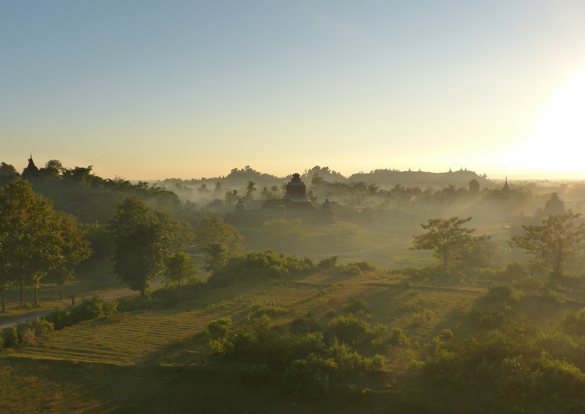 Mist rises over the fields and stupas of Mrauk-U at dawn, silhouetted temples visible along the horizon.