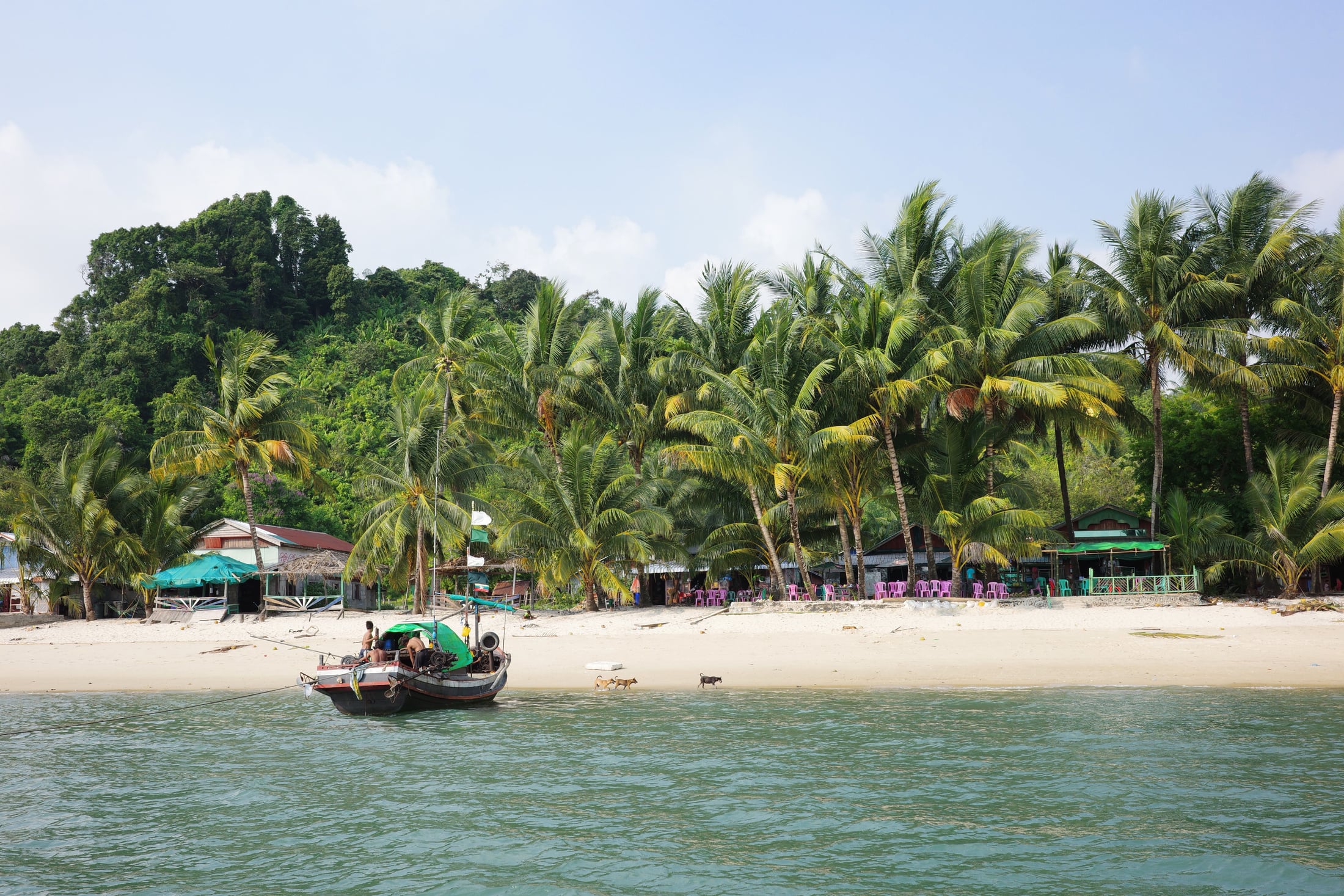 A traditional wooden boat rests on a pale sand beach fringed with palm trees at Myeik, with a small coastal settlement visible among the trees.