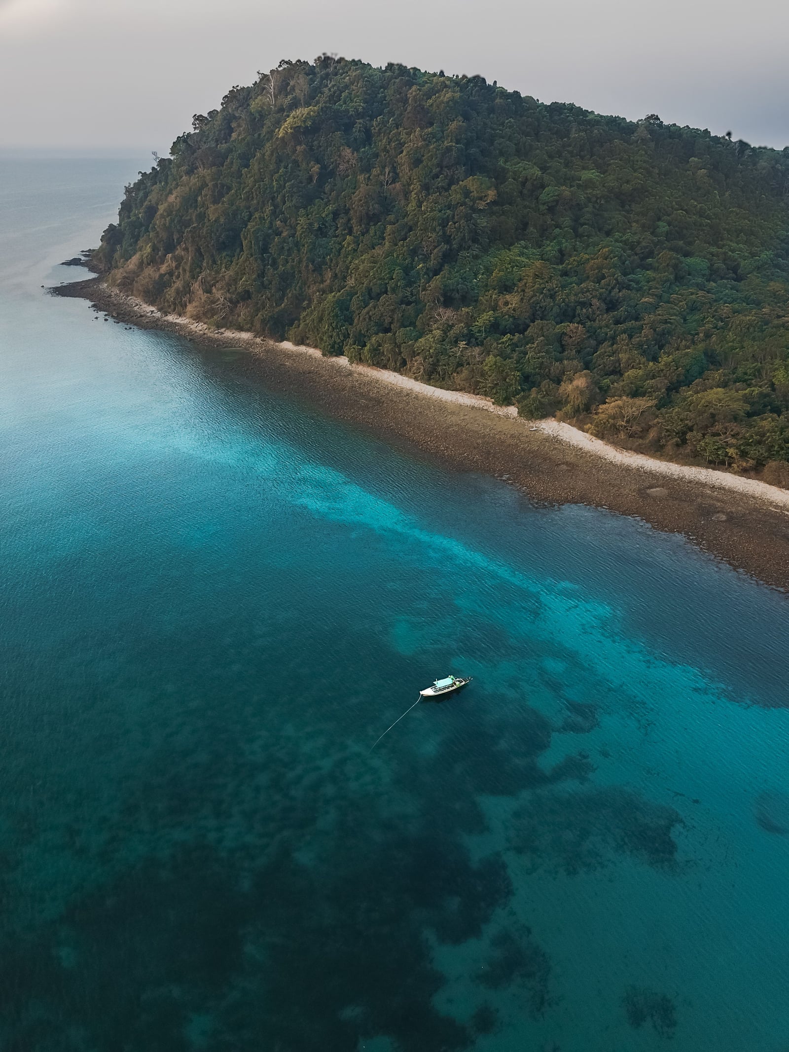 A small boat is anchored over a shallow reef near the tropical coastline of Dawei, the water shading from pale turquoise to deep blue.