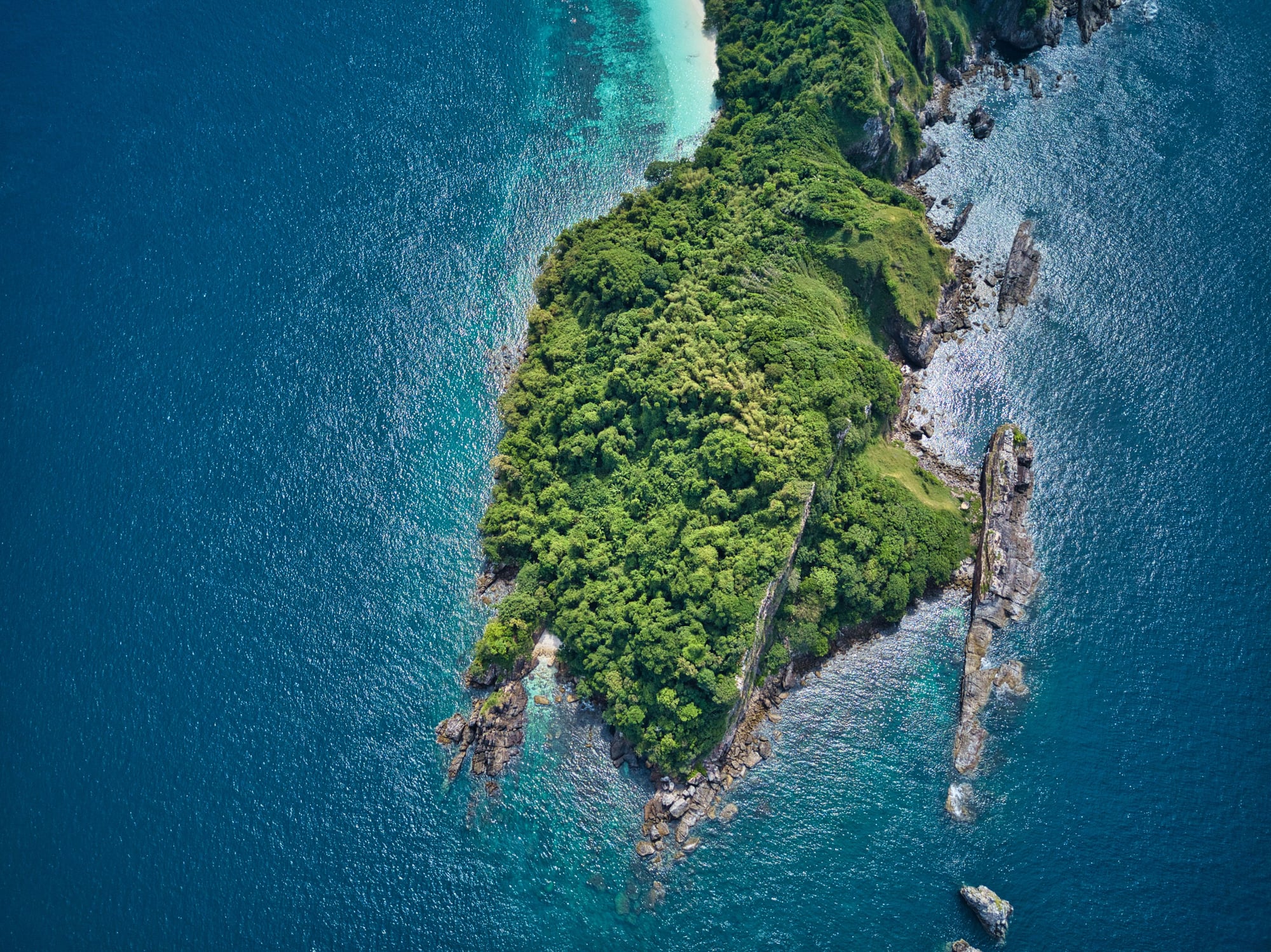 An aerial view of a forested island near Kawthoung, its rocky headland meeting turquoise reef water and a pale sand beach.