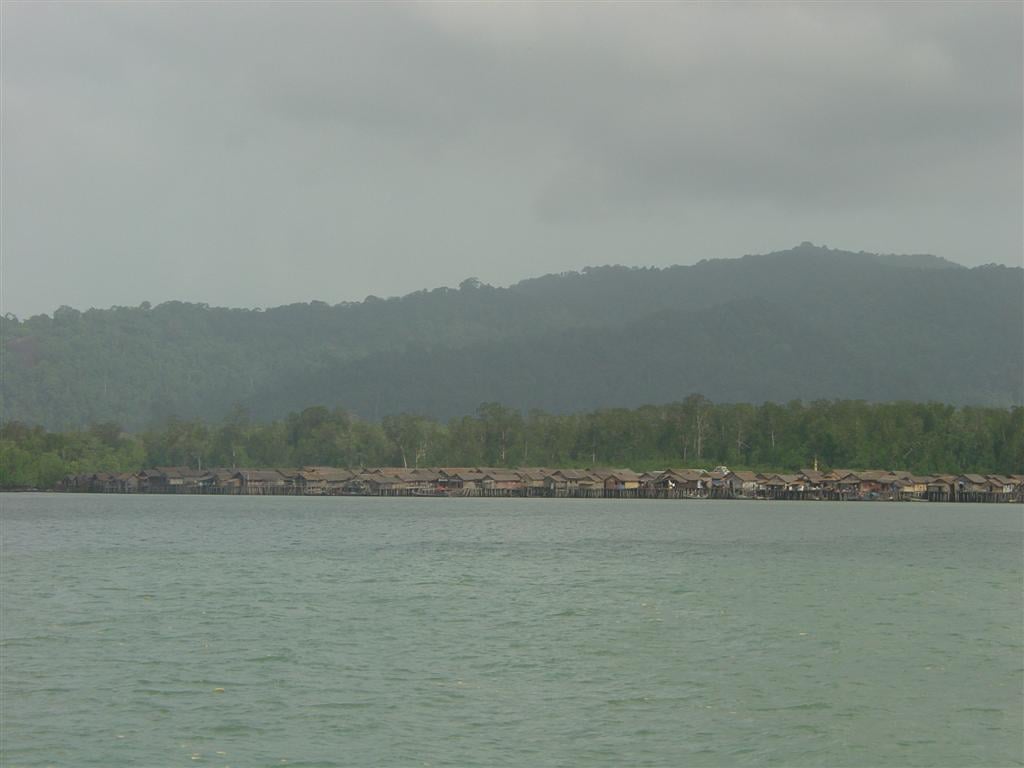 A stilt village lines the shore of an island in the Myeik Archipelago, backed by forested hills under a grey sky.