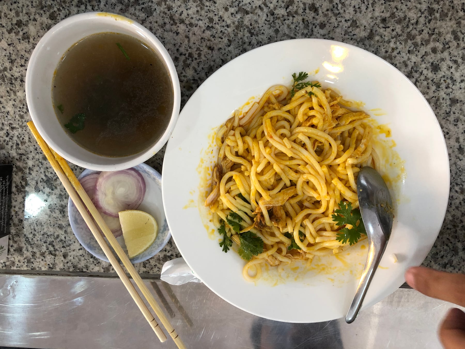 A plate of nan gyi thoke, thick rice noodles tossed with chicken curry, served with a side of soup, onion and lime.