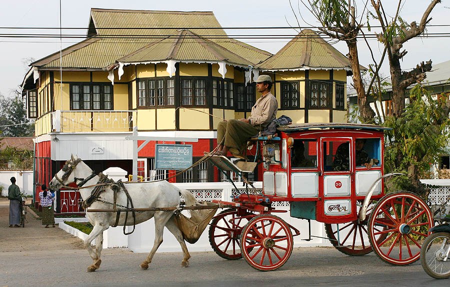 A white horse pulls a red-wheeled carriage past a yellow colonial-era building in Pyin Oo Lwin, with the driver seated on top.