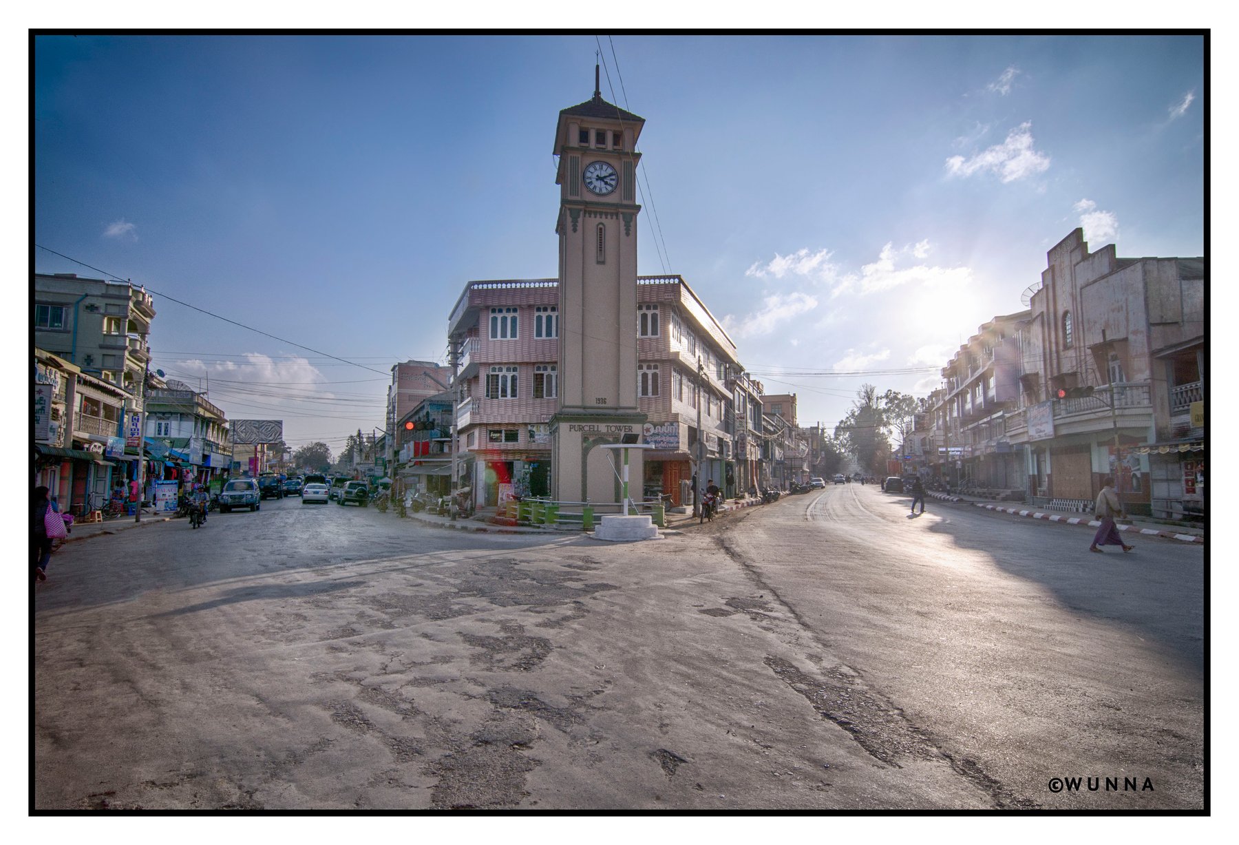 The Purcell Clock Tower stands at a sunlit street intersection in Pyin Oo Lwin, with shops and colonial-era buildings lining the road on either side.