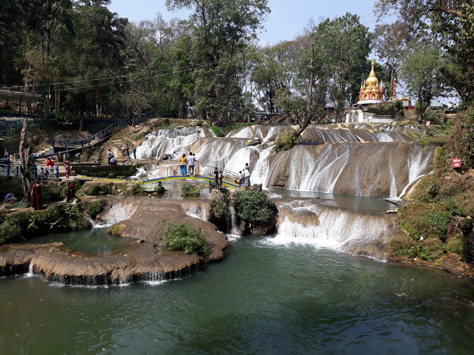 Water cascades over several tiers of rock at Pwe Kauk Falls near Pyin Oo Lwin, with a footbridge, visitors, and a golden pagoda spire visible among the trees.