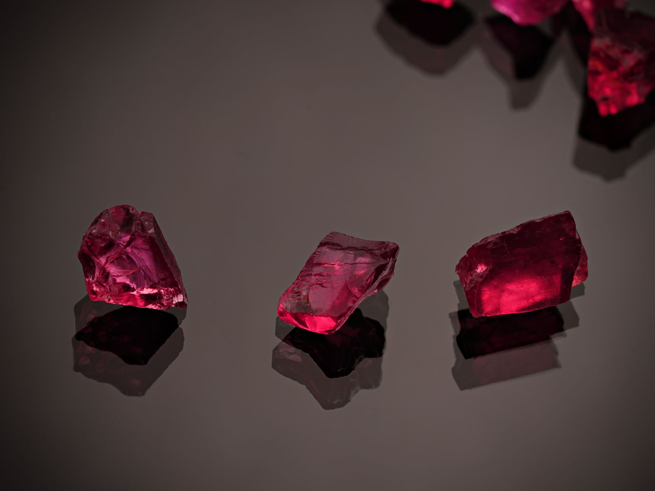 Three raw, glittering red ruby crystals reflected on a polished black surface.