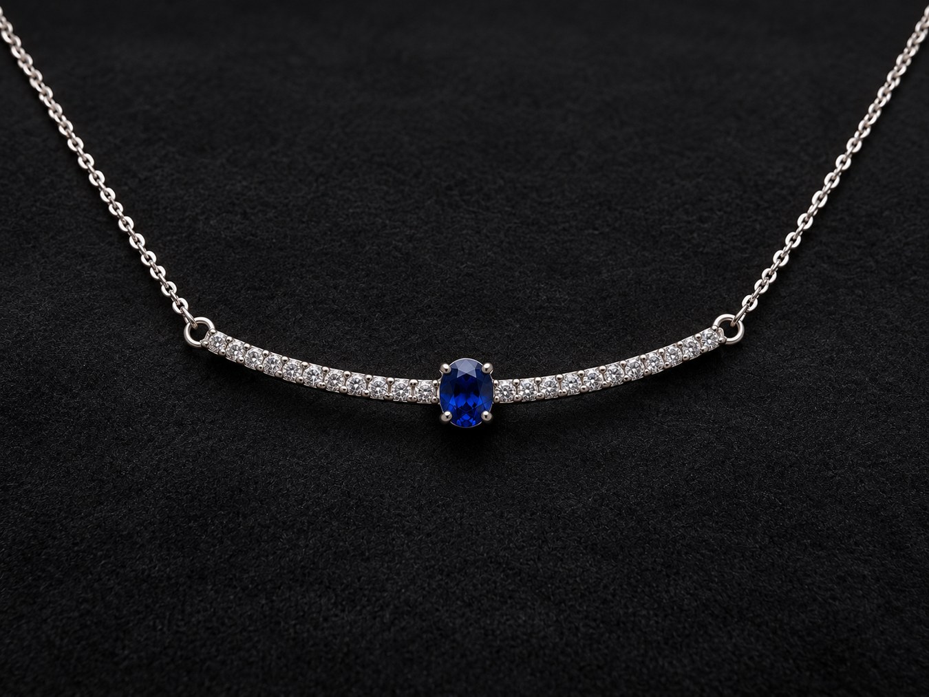 A white gold necklace with a diamond-set curved bar and a central oval blue sapphire, resting on black fabric.