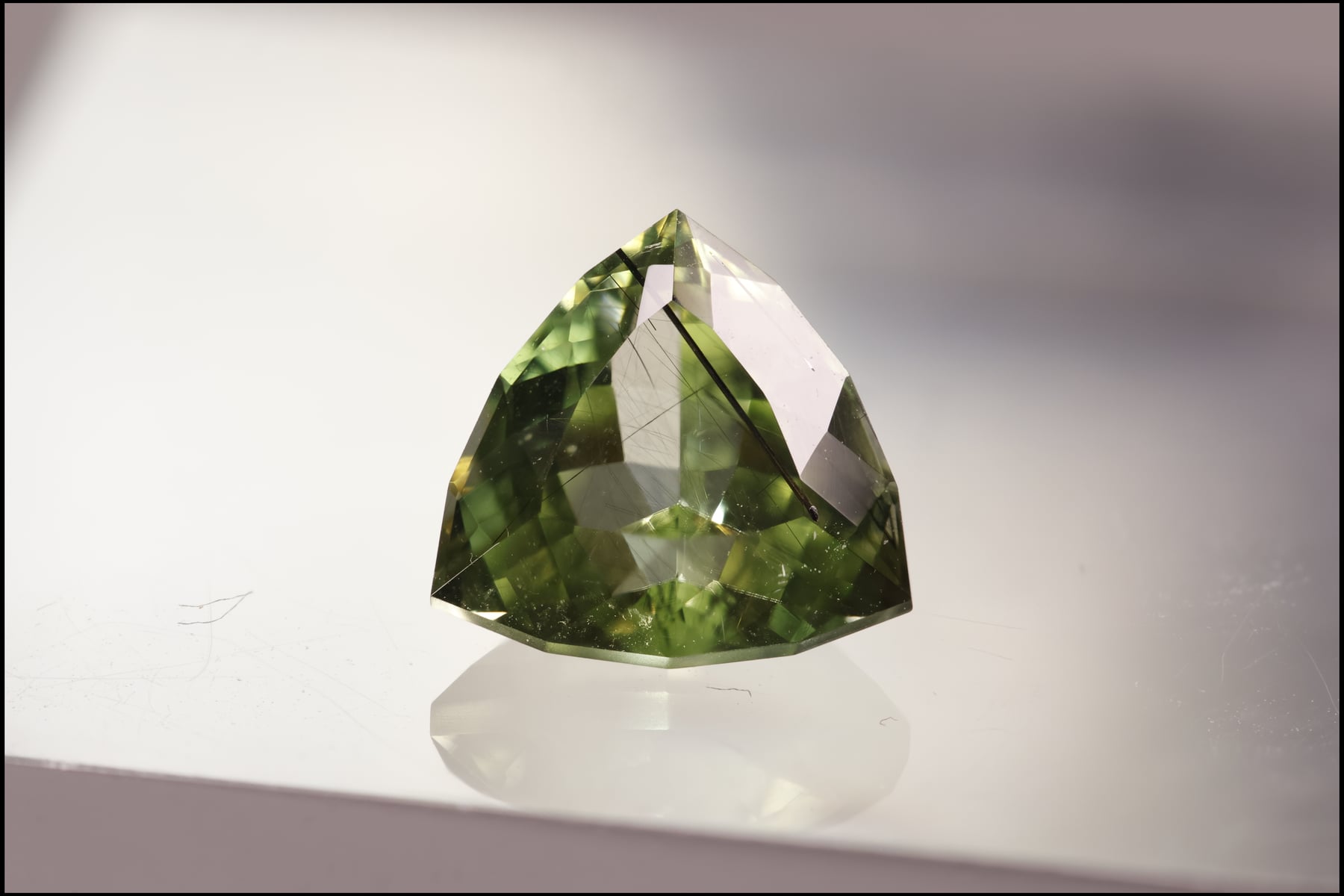 A trillion-cut green peridot gemstone with visible needle-like inclusions, photographed against a soft grey background.