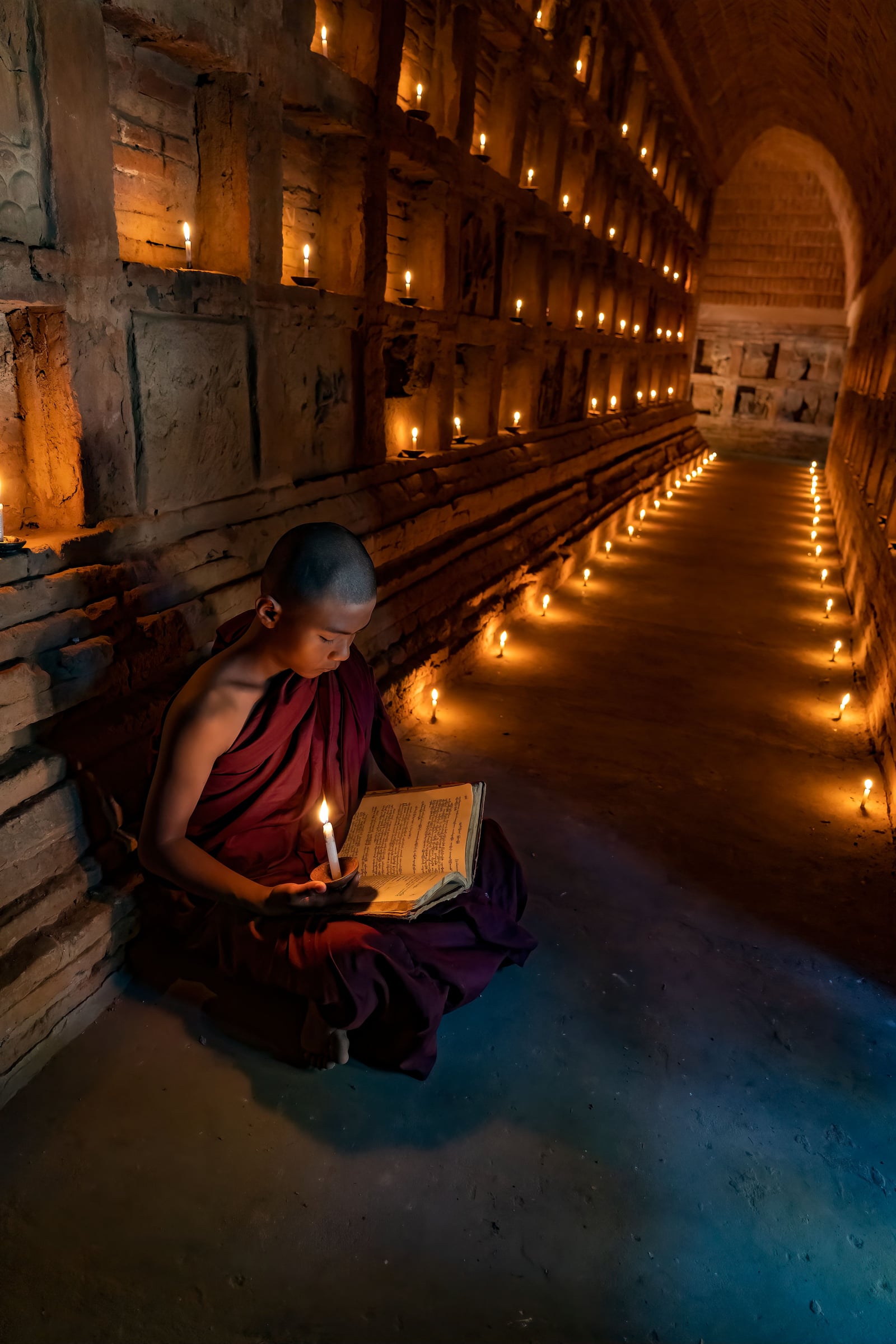 A young novice monk sits reading scripture by candlelight in a temple corridor lined with lit candles.