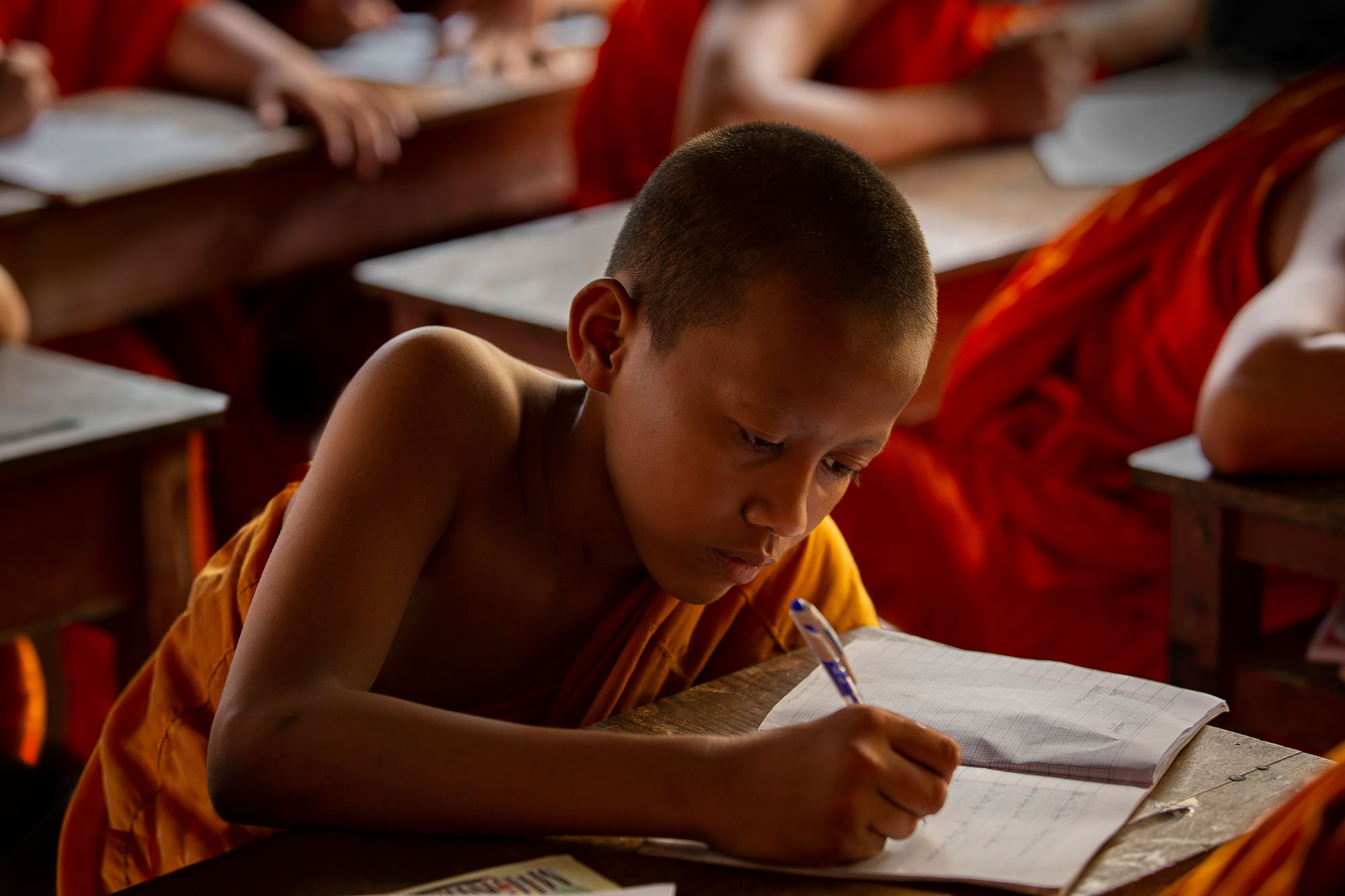 A young novice monk in orange robes writes carefully in a notebook at a classroom desk.