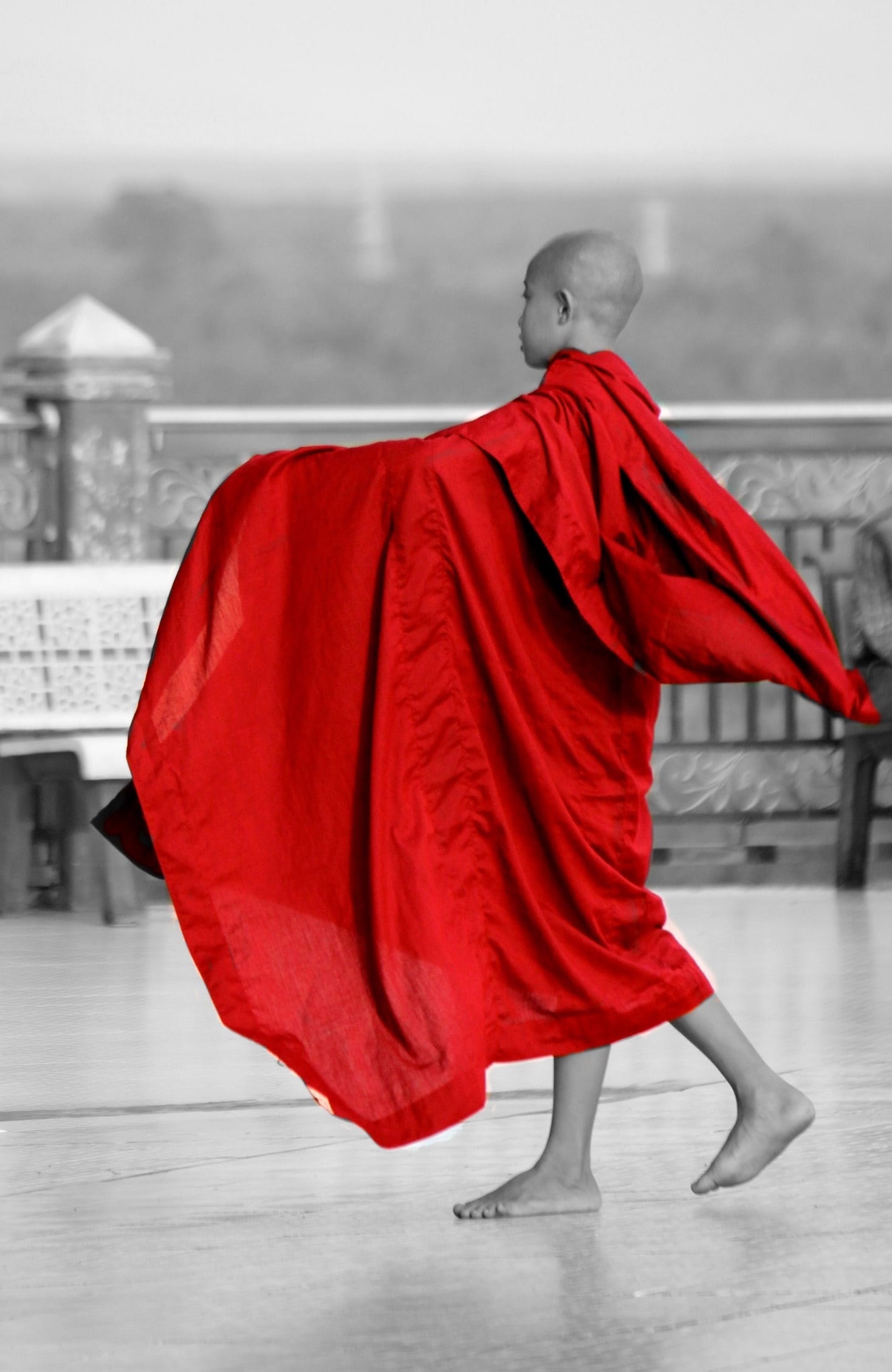 A young novice monk's red robe billows in the wind as he walks across a temple terrace.