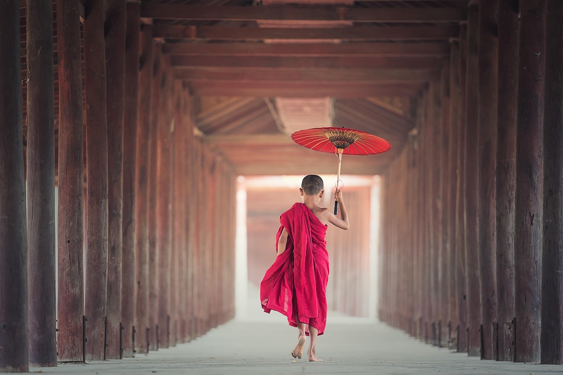 A young novice monk in a red robe holds a red parasol as he walks down a wooden temple corridor.