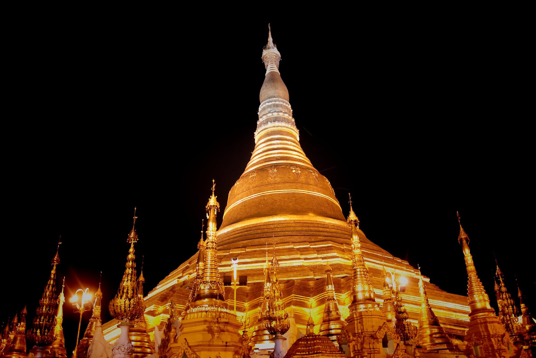 A close, low-angle view of Shwedagon's golden stupa at night, its tiered terraces and slender spire lit against a black sky, with the silver hti umbrella crown at the very top.
