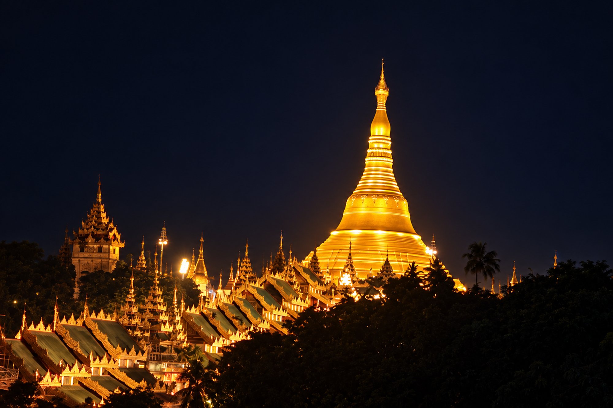 Shwedagon Pagoda's golden stupa rises above the green-roofed pavilions of its platform at night, with a palm tree silhouette and a smaller spired hall to one side.