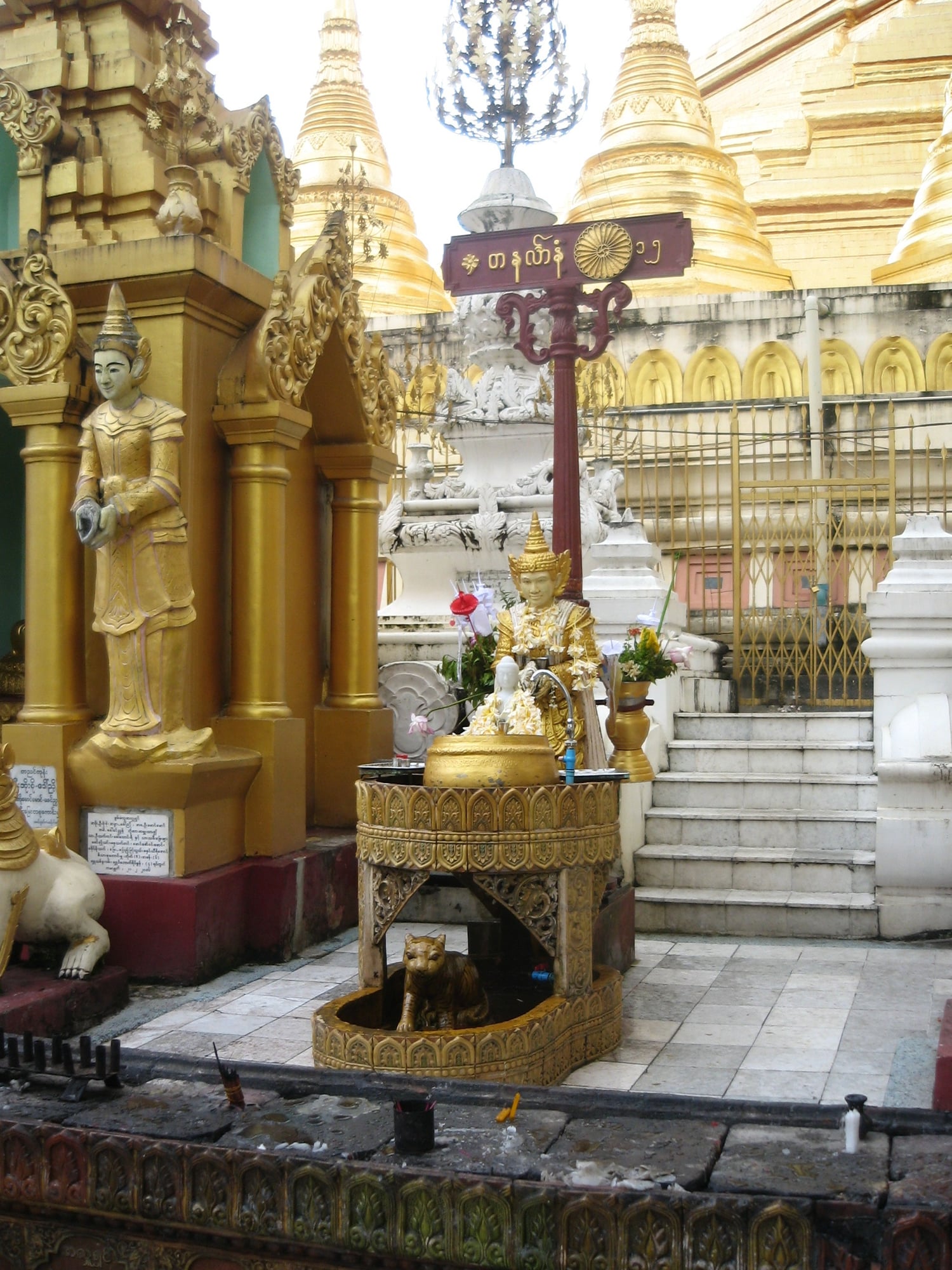 A planetary post shrine at Shwedagon, with a gilded guardian figure and a small basin where devotees pour water over a Buddha image.