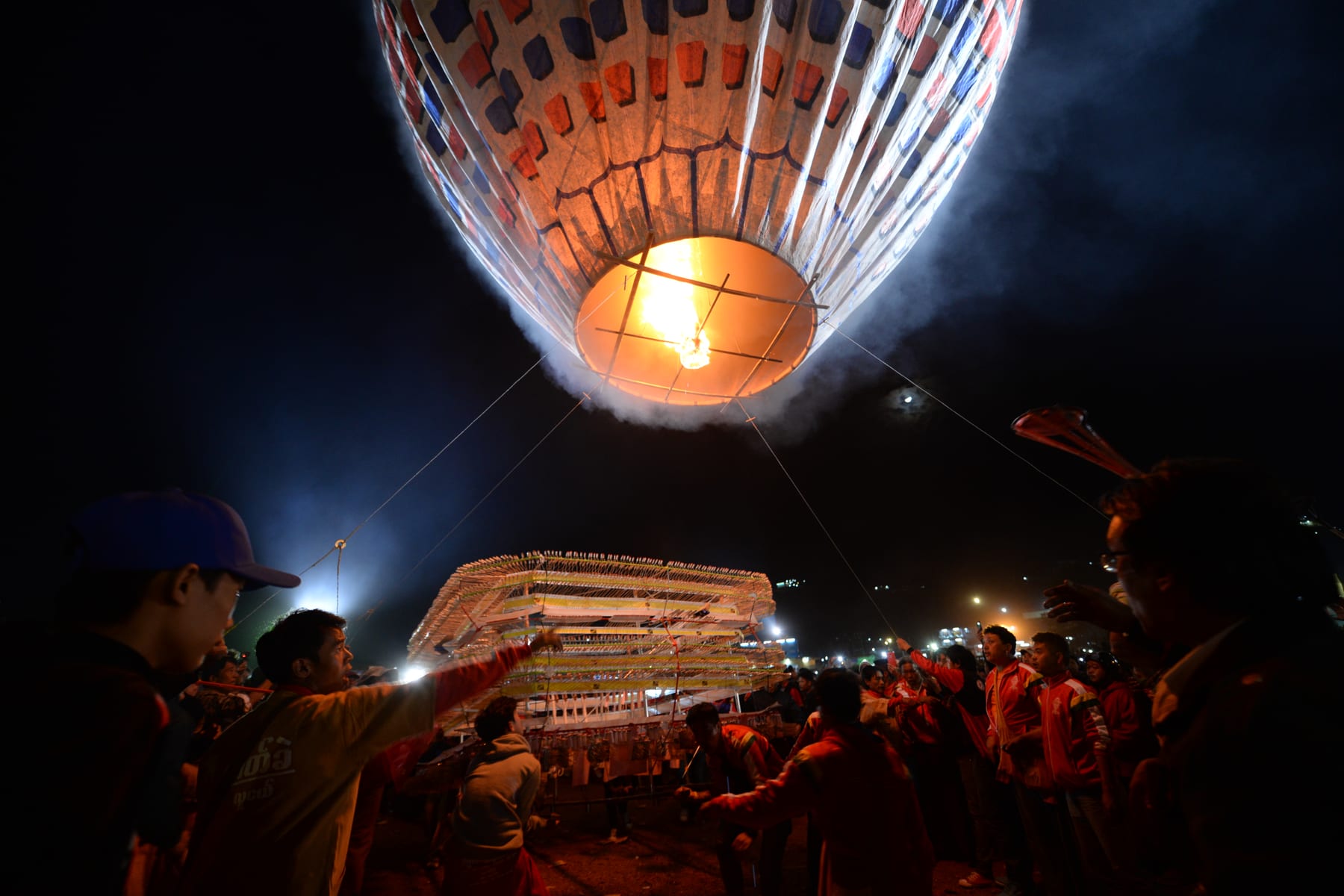 A crowd helps steady a hot air balloon rigged with a tiered firework frame as it is prepared for a night launch, its paper envelope glowing from the flame inside.