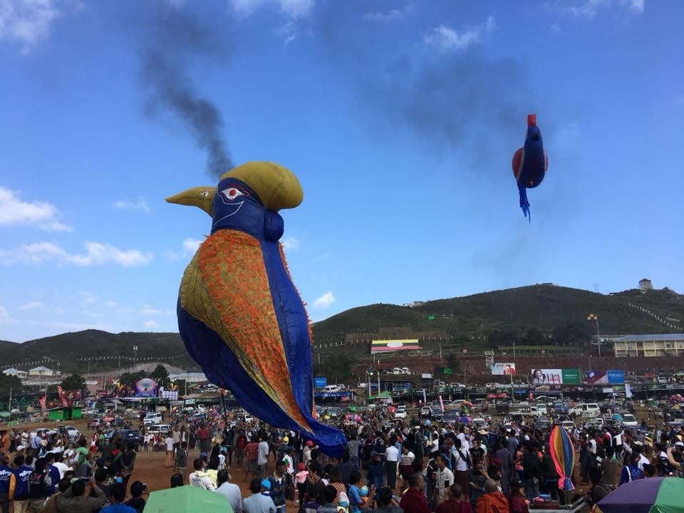 A giant paper balloon shaped like an Auk-Chin bird, painted in blue and orange, rises above a crowd with the Shan hills behind it.