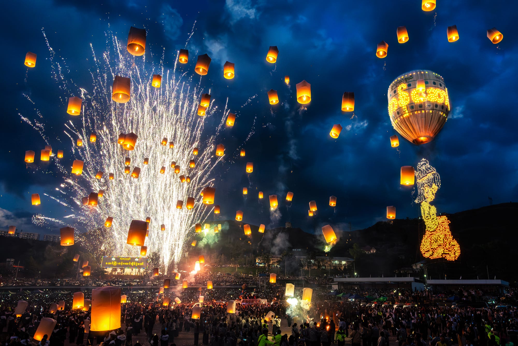 Fireworks explode beside dozens of glowing paper lanterns and a giant illuminated hot air balloon rising into the night sky at the Taunggyi festival.