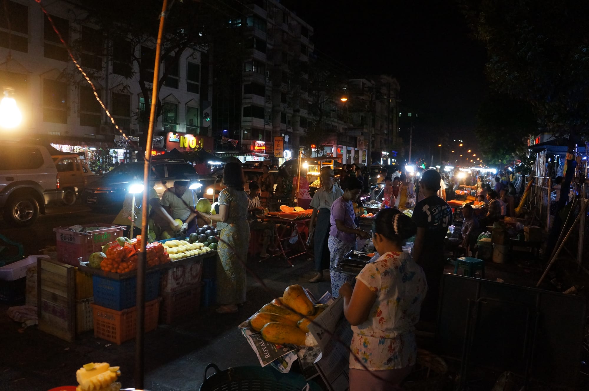 A bustling night street food market in Yangon, with fruit stalls lit by bare bulbs and crowds of shoppers.