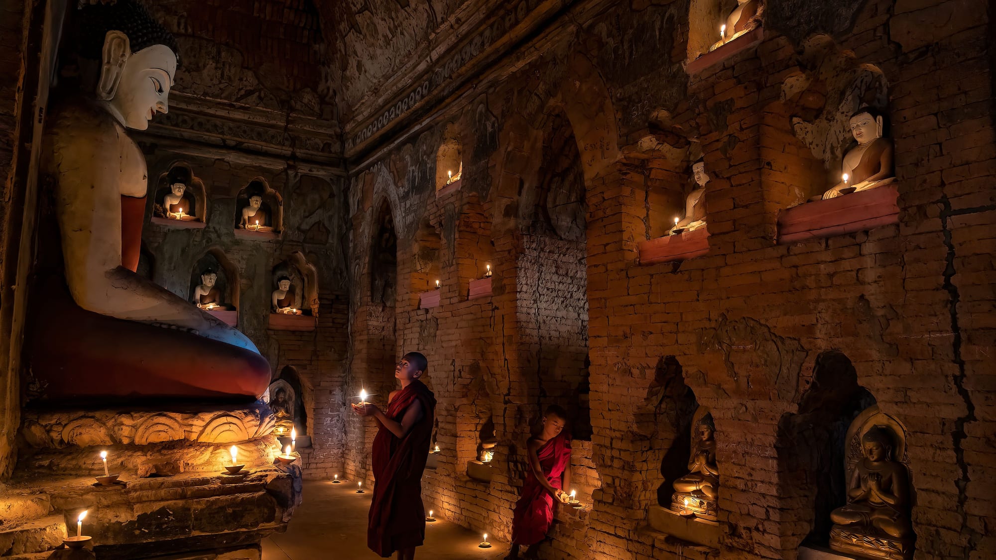 Novice monks light candles among rows of ancient Buddha statues inside a candlelit Bagan temple.
