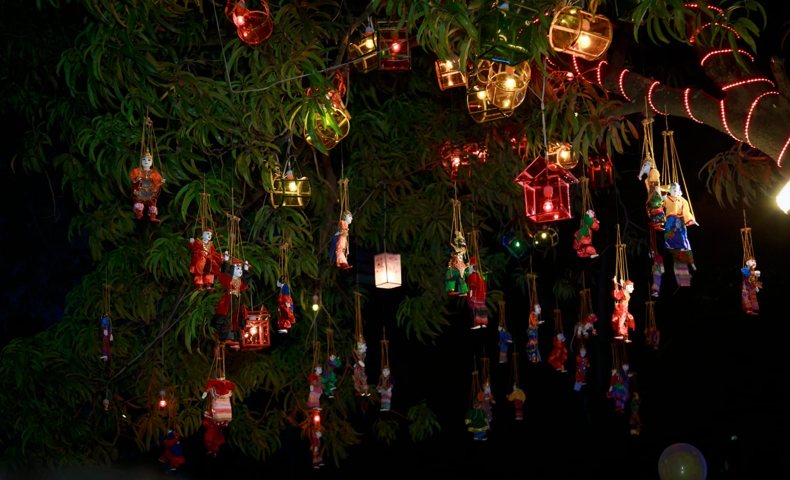 Colourful lanterns and traditional marionette puppets hang from the branches of a tree, lit up at night.