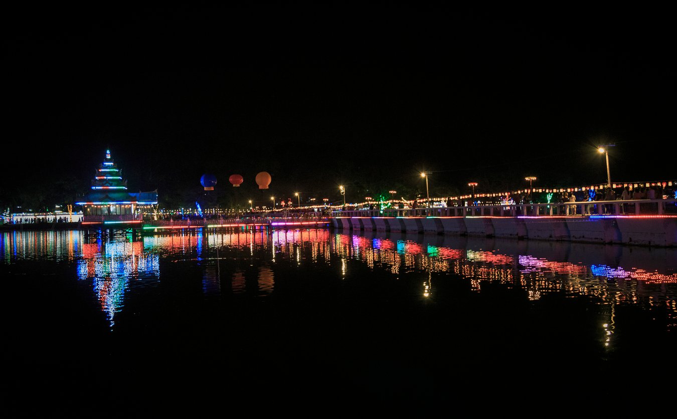 A lit pagoda and a lantern-strung bridge reflect in the still water of a lake at night during Thadingyut.