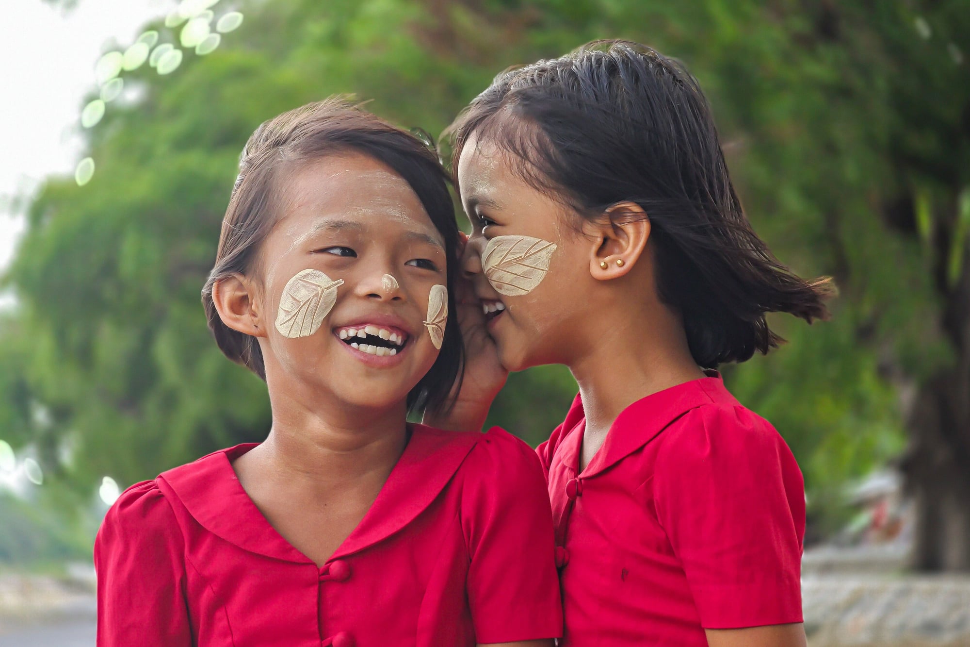 Two laughing girls in red dresses lean close together, each wearing a leaf-patterned design of thanaka paste on her cheeks, in Amarapura.