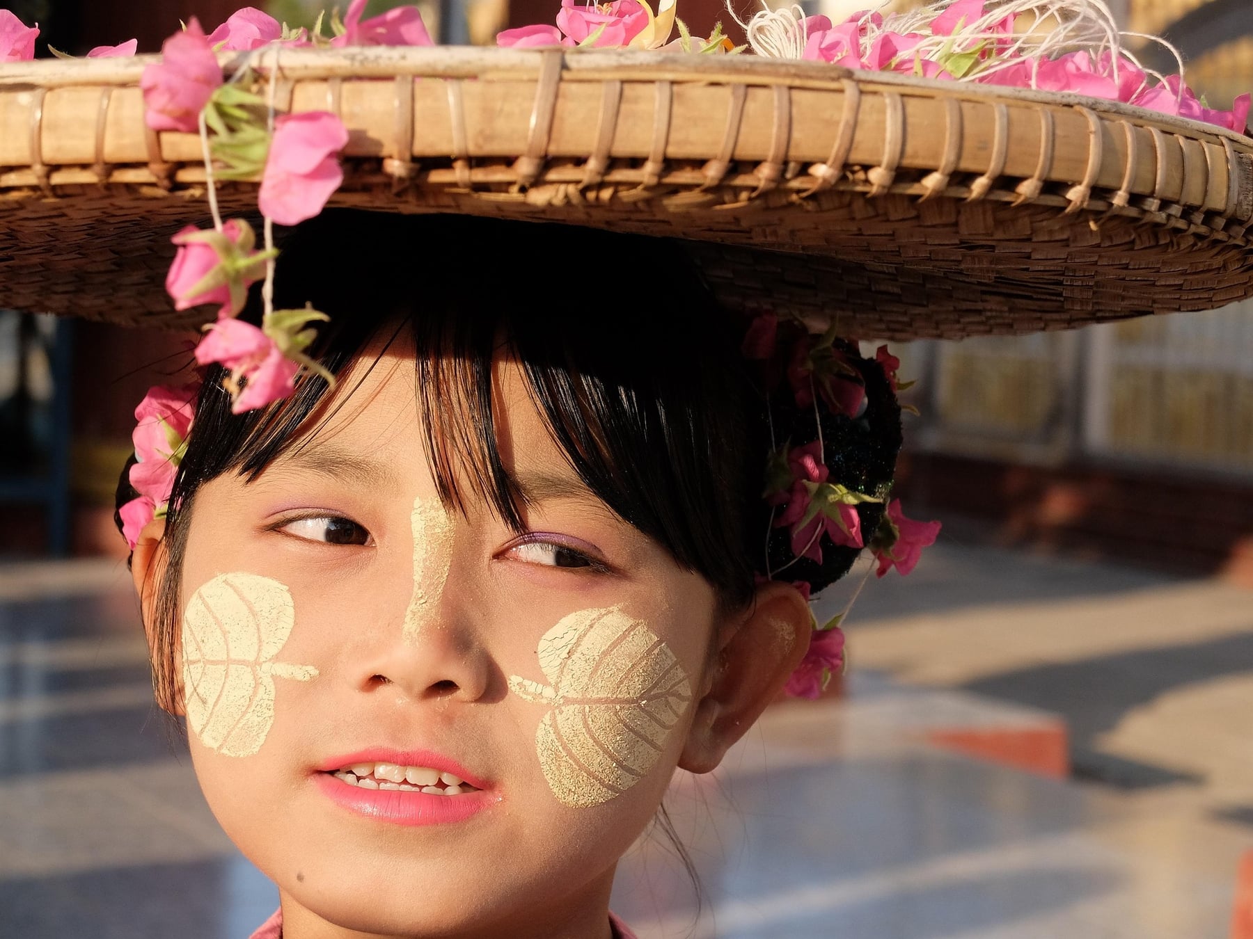 A girl wearing a wide flower-decorated hat, with a leaf-patterned design of thanaka paste freshly applied to her cheeks.