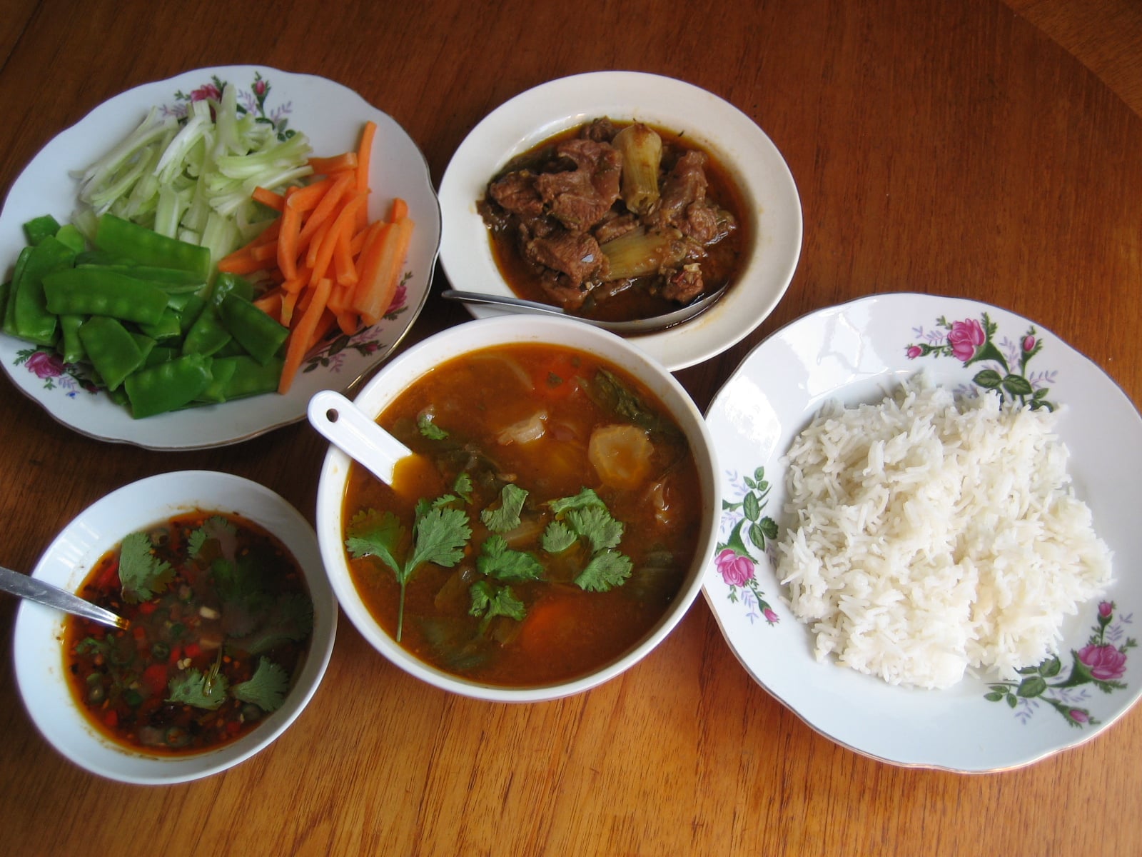 An overhead view of a Burmese home meal, with rice, beef curry, sour soup, raw vegetables and a bowl of ngapi yay dip.