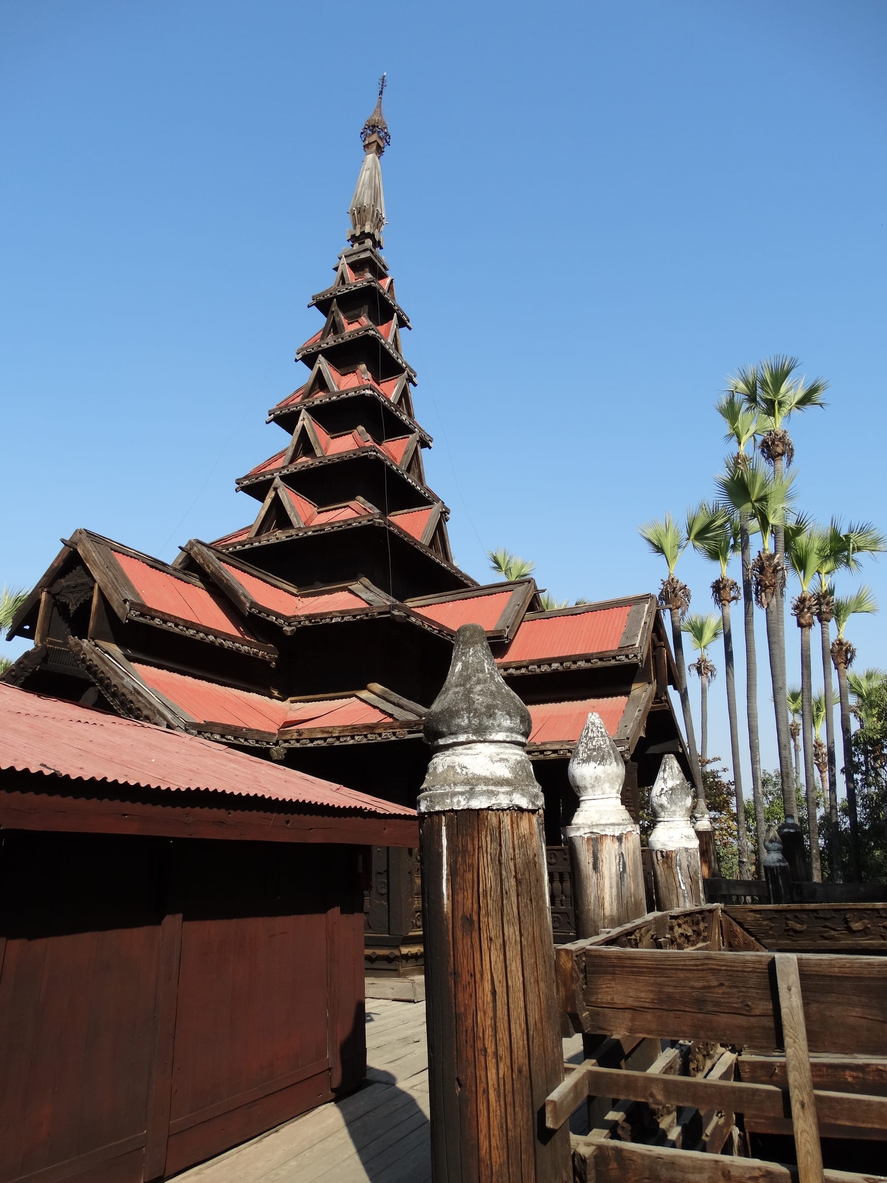 The tiered teak roof of Bagaya Monastery in Inwa rising against a clear blue sky, framed by palm trees.