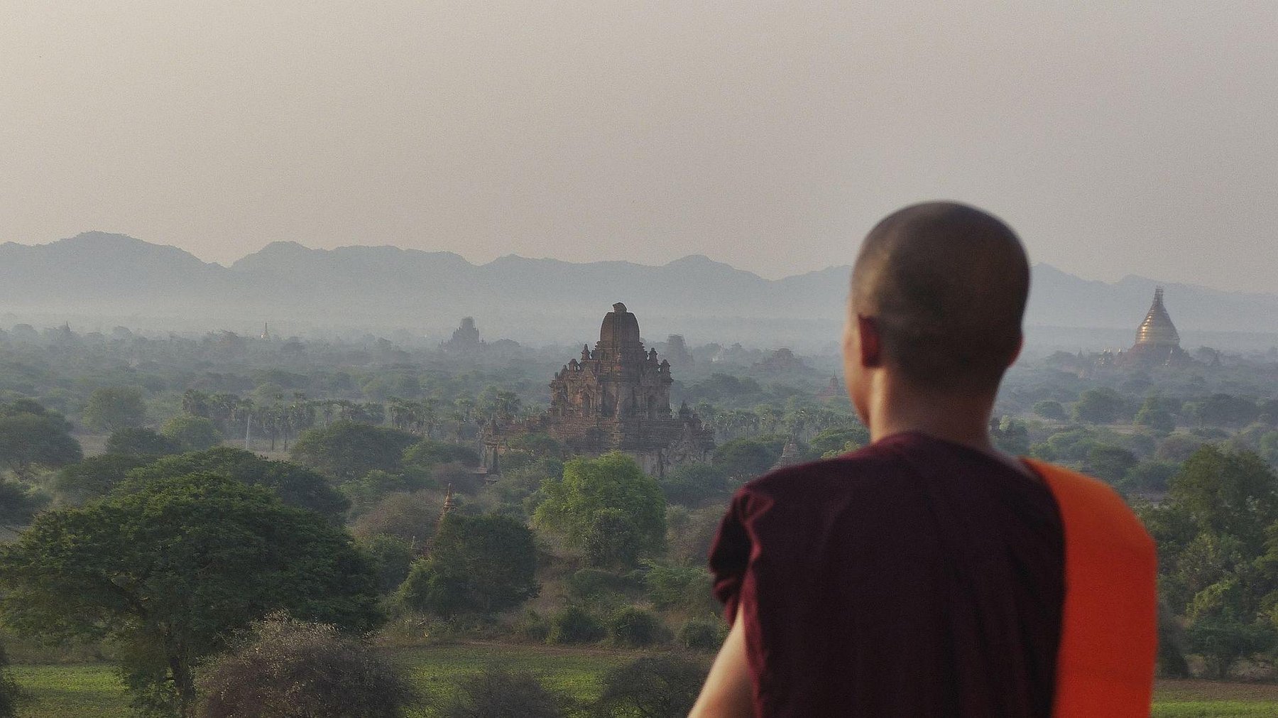 A young monk looks out over the temple-studded plain of Bagan at dawn, mist rising among the trees and stupas.