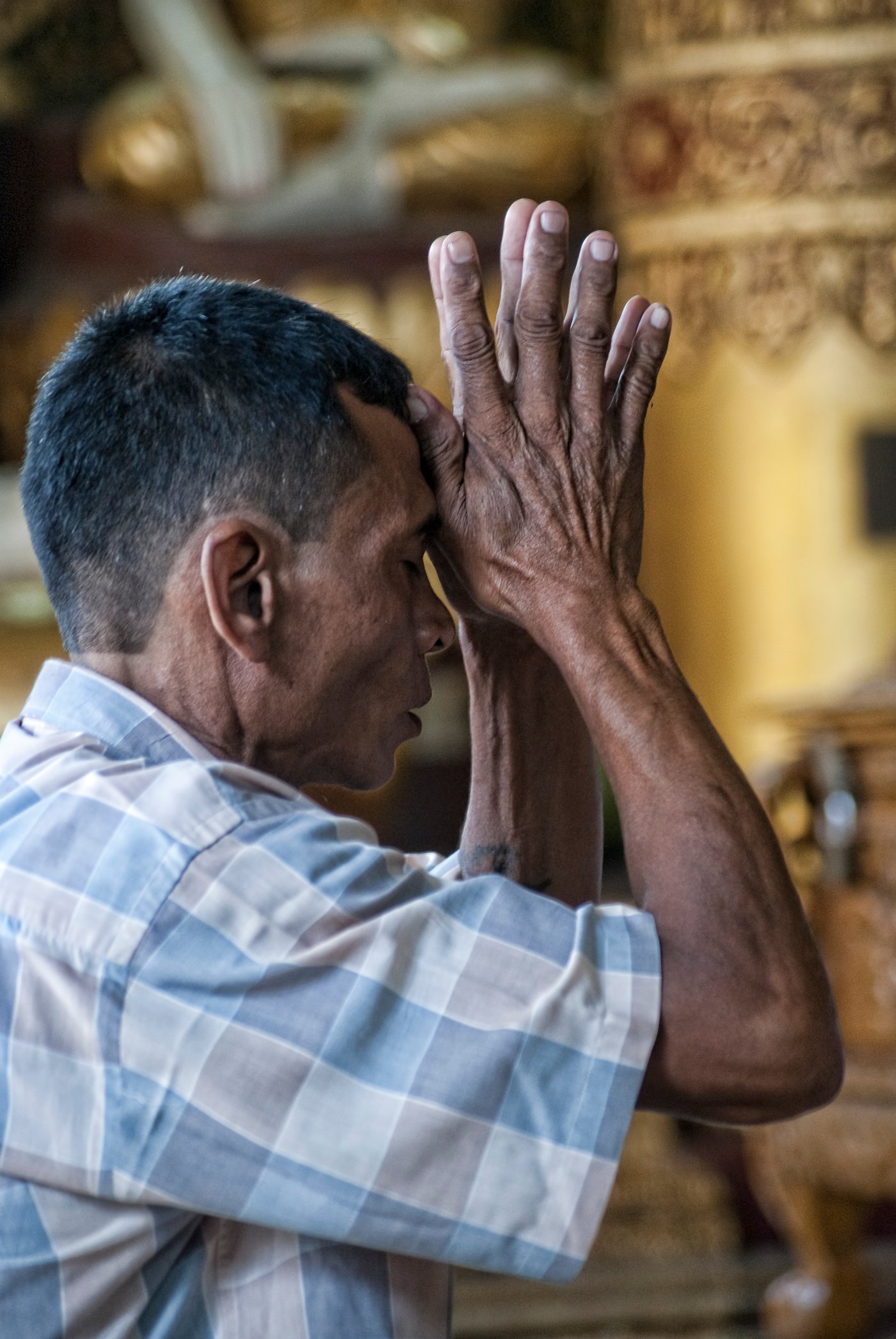 A man presses his hands together in prayer at his forehead, eyes closed, inside a gilded temple hall.
