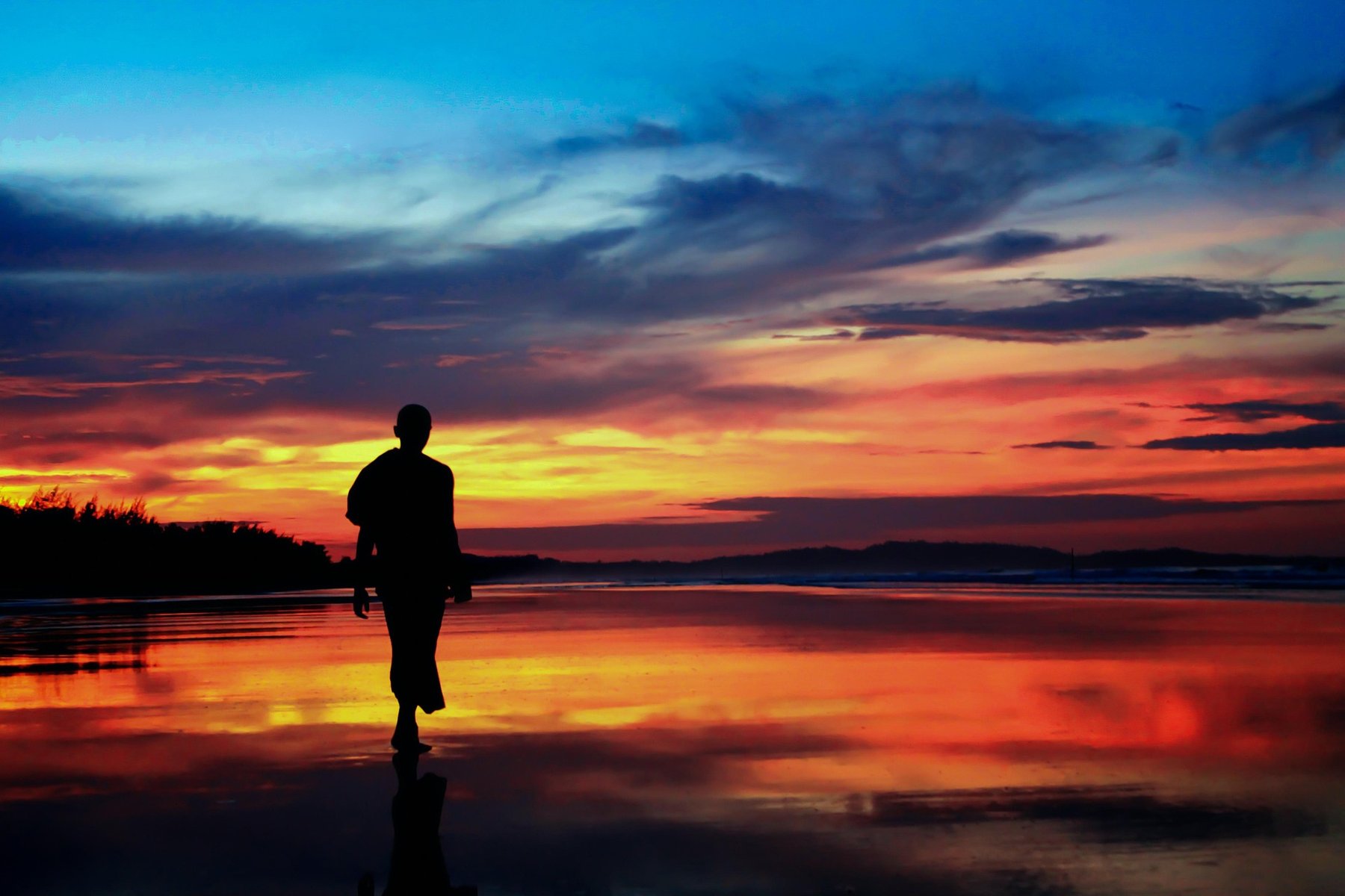 The silhouette of a monk walking along a reflective tidal beach at dusk, the sky glowing orange and red.