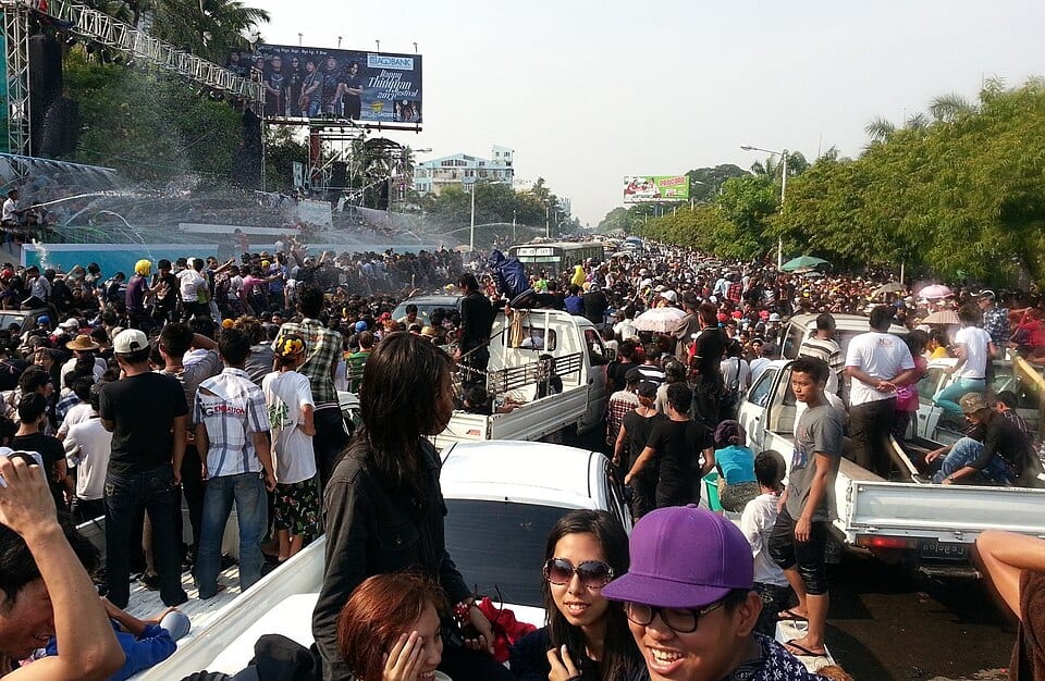 A dense crowd fills a city street during Thingyan, packed onto trucks and drenched with water from a stage-mounted hose.