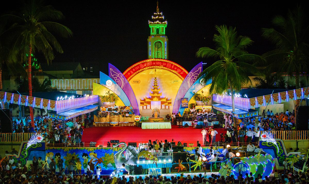 A brightly lit central pavilion and music stage, decorated with a float and palm trees, draws a packed crowd at night during Mandalay's Thingyan festival.