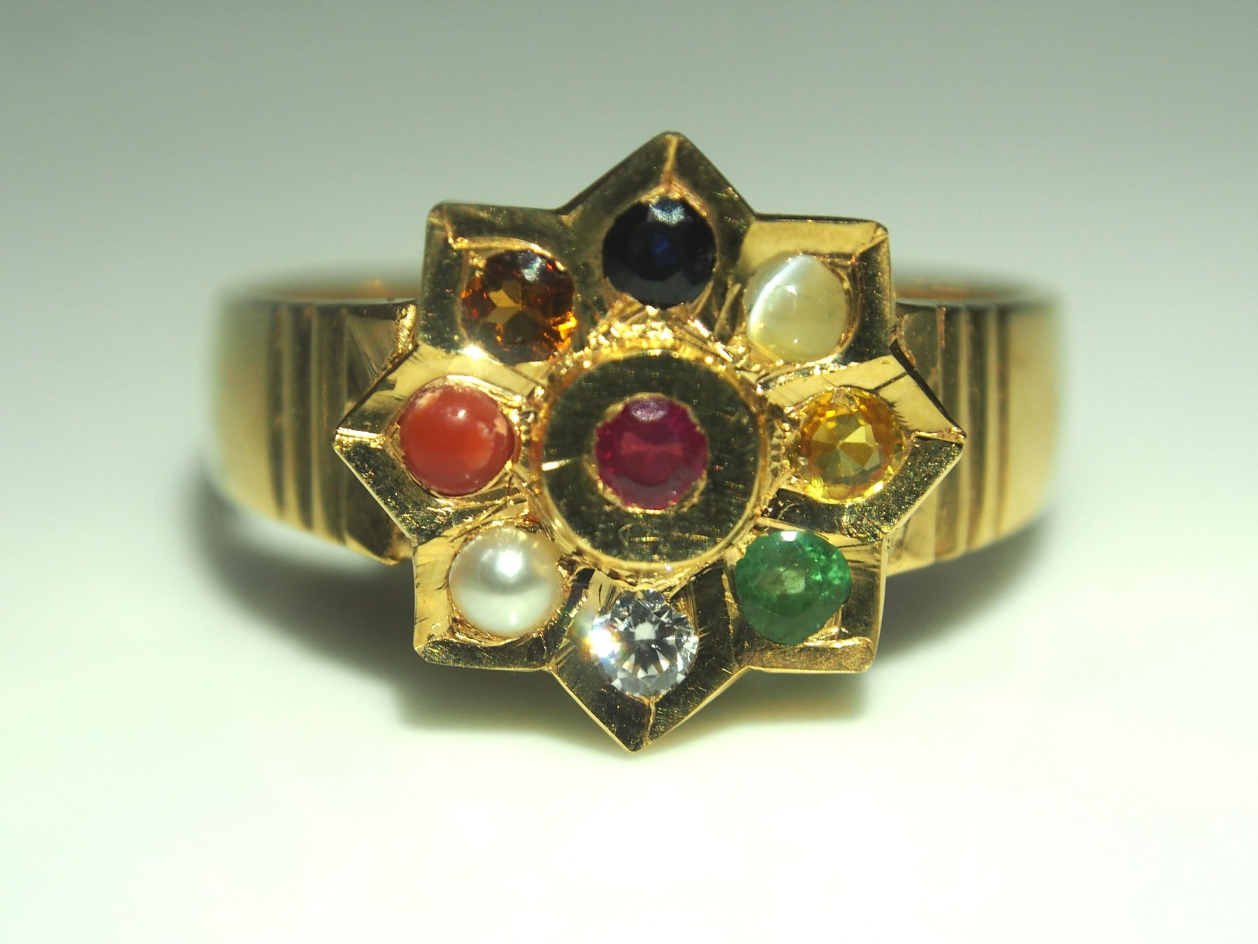 A gold ring set with nine different gemstones arranged around a central red stone, in the nawarat or navaratna pattern.