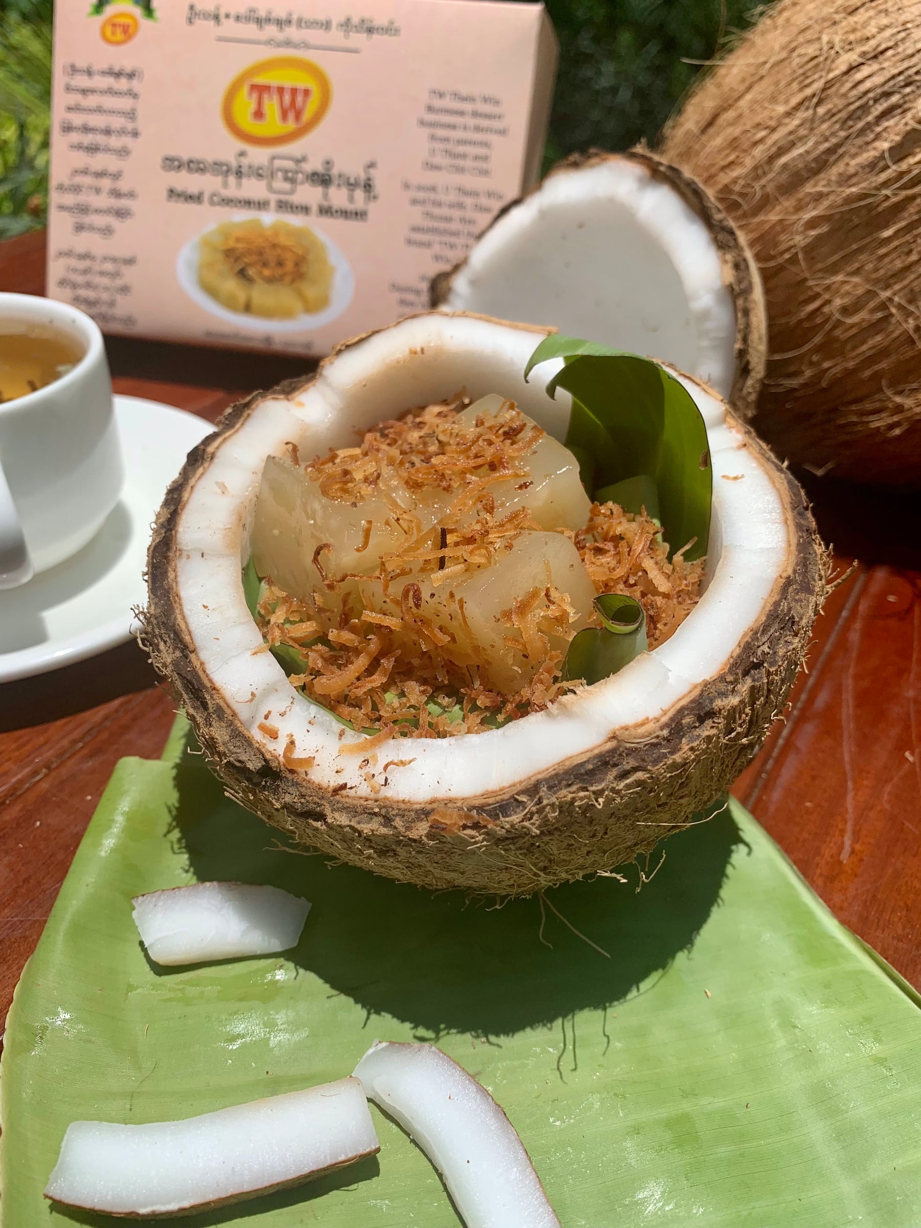 Htoe mont, a glutinous rice sweet topped with fried coconut, served inside a coconut shell alongside a cup of tea.